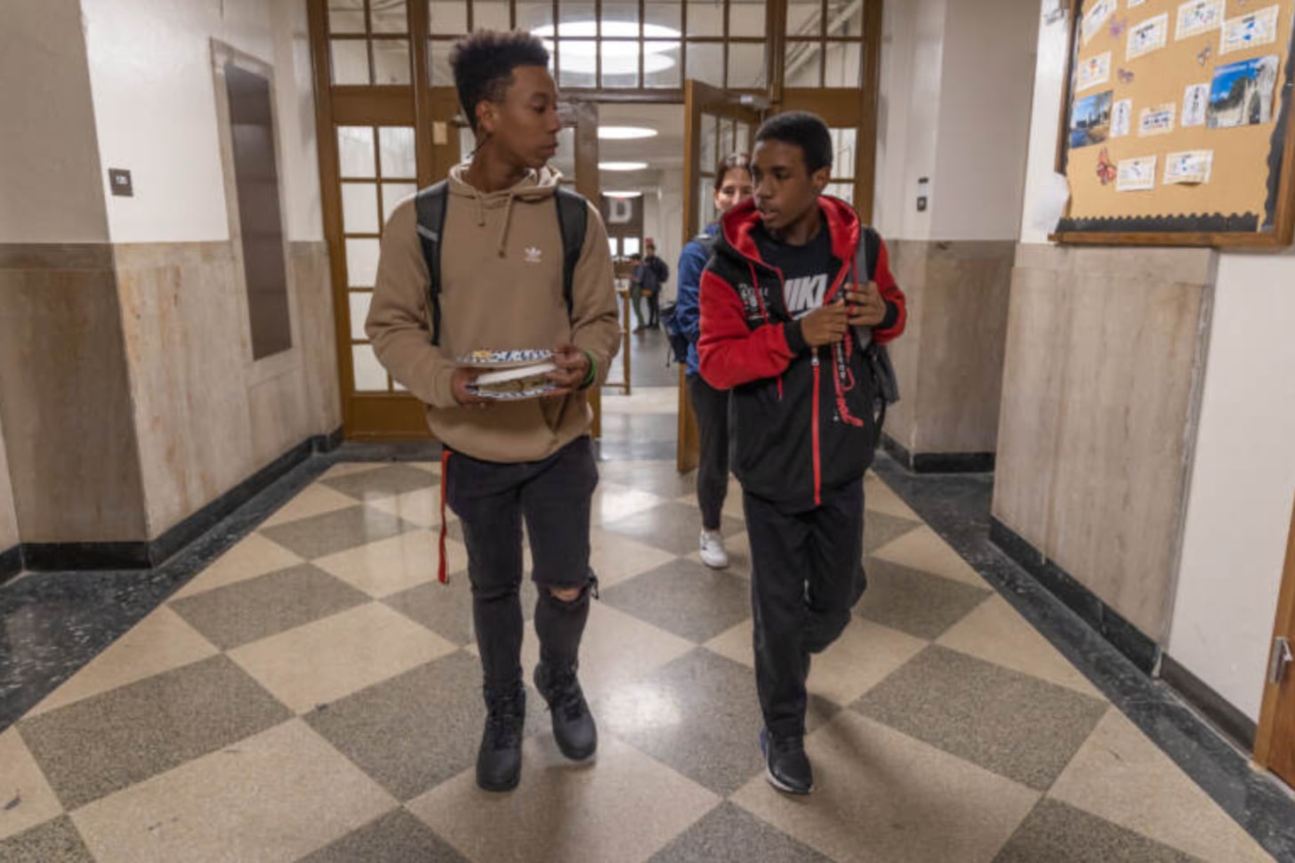 A student wearing a brown hooded sweater and black jeans and black shoes walks next to another student with short hair and a red and black sweater.