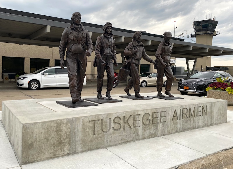 A moment of four people stand on top of a concrete platform with the words "Tuskegee Airmen" carved on the side with a car and an airport in the background.