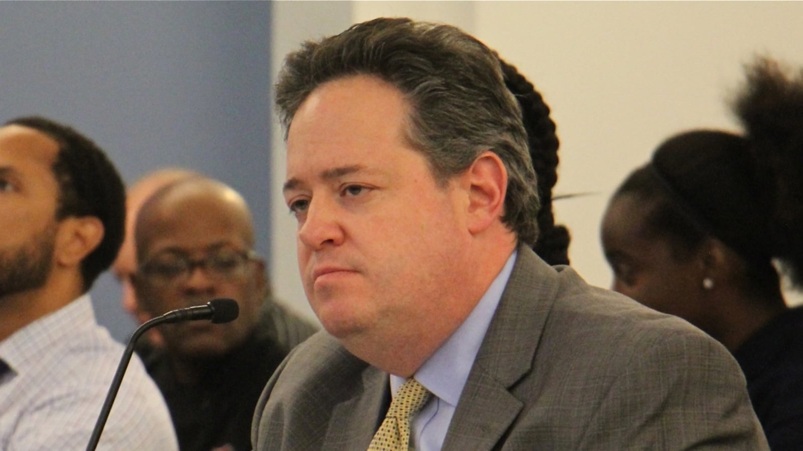 A man wearing a dark suit jacket, blue shirt, and tie looks straight ahead with a microphone in front of him.
