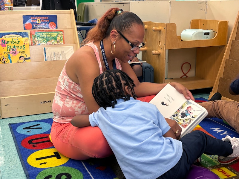 An adult wearing a pink outfit sits and reads to a young student on a colorful rug in a classroom.