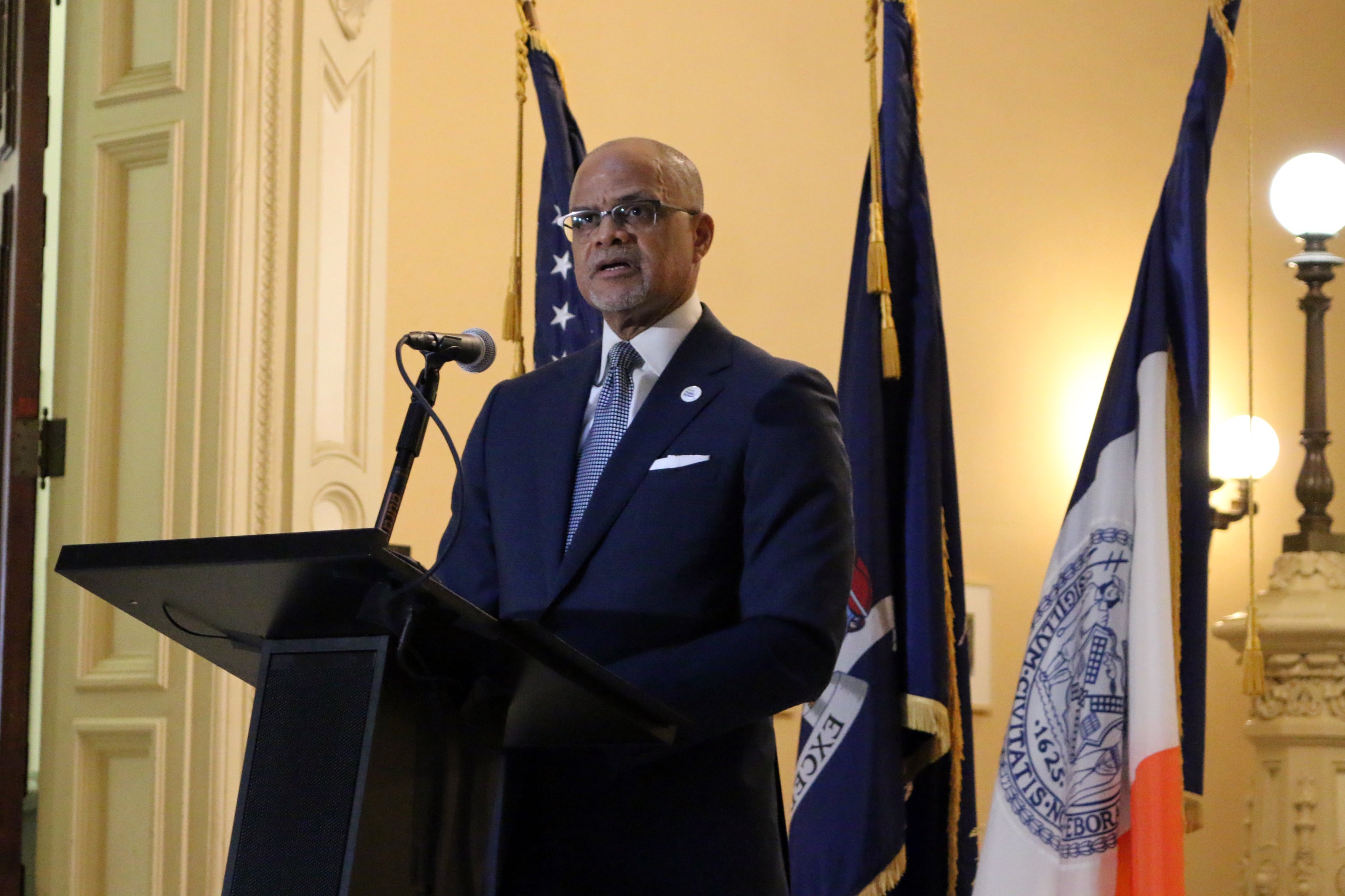 A man wearing glasses and a dark suit speaks from a podium with three flags in the background.