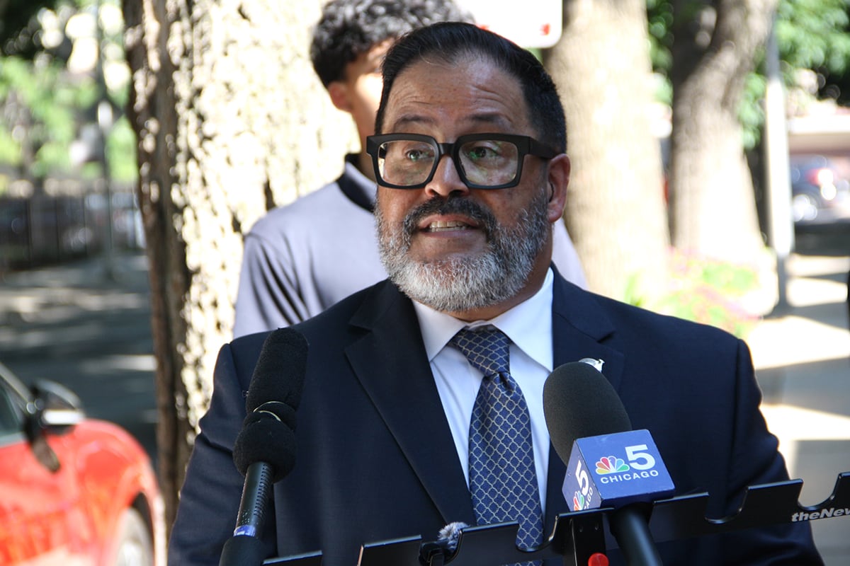 A man in a dark suit jacket and glasses stands in front of microphones.