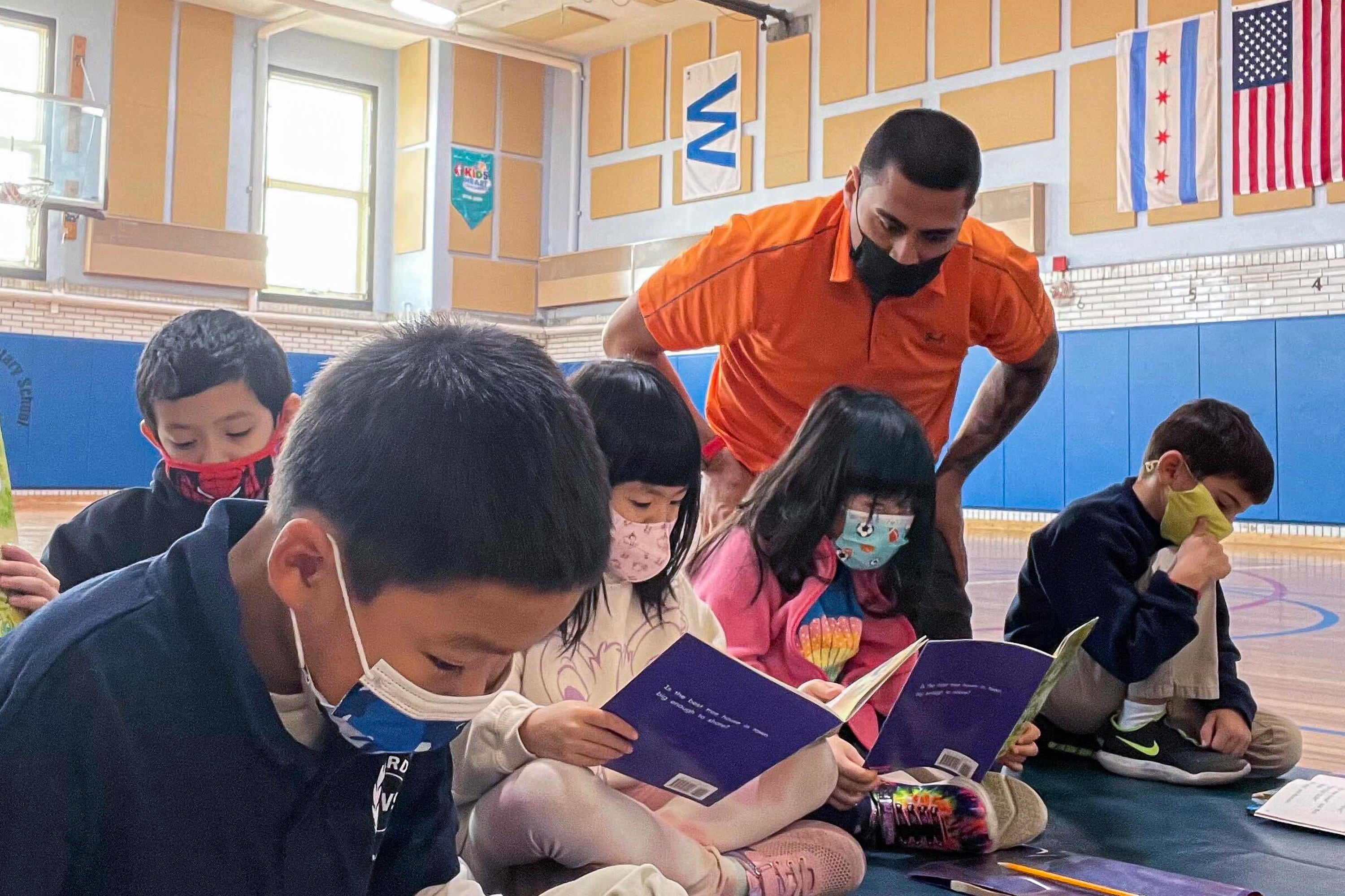 A teacher wearing an orange polo looks over the shoulders of several young students, who are reading in a school gymnasium and wearing protective masks.