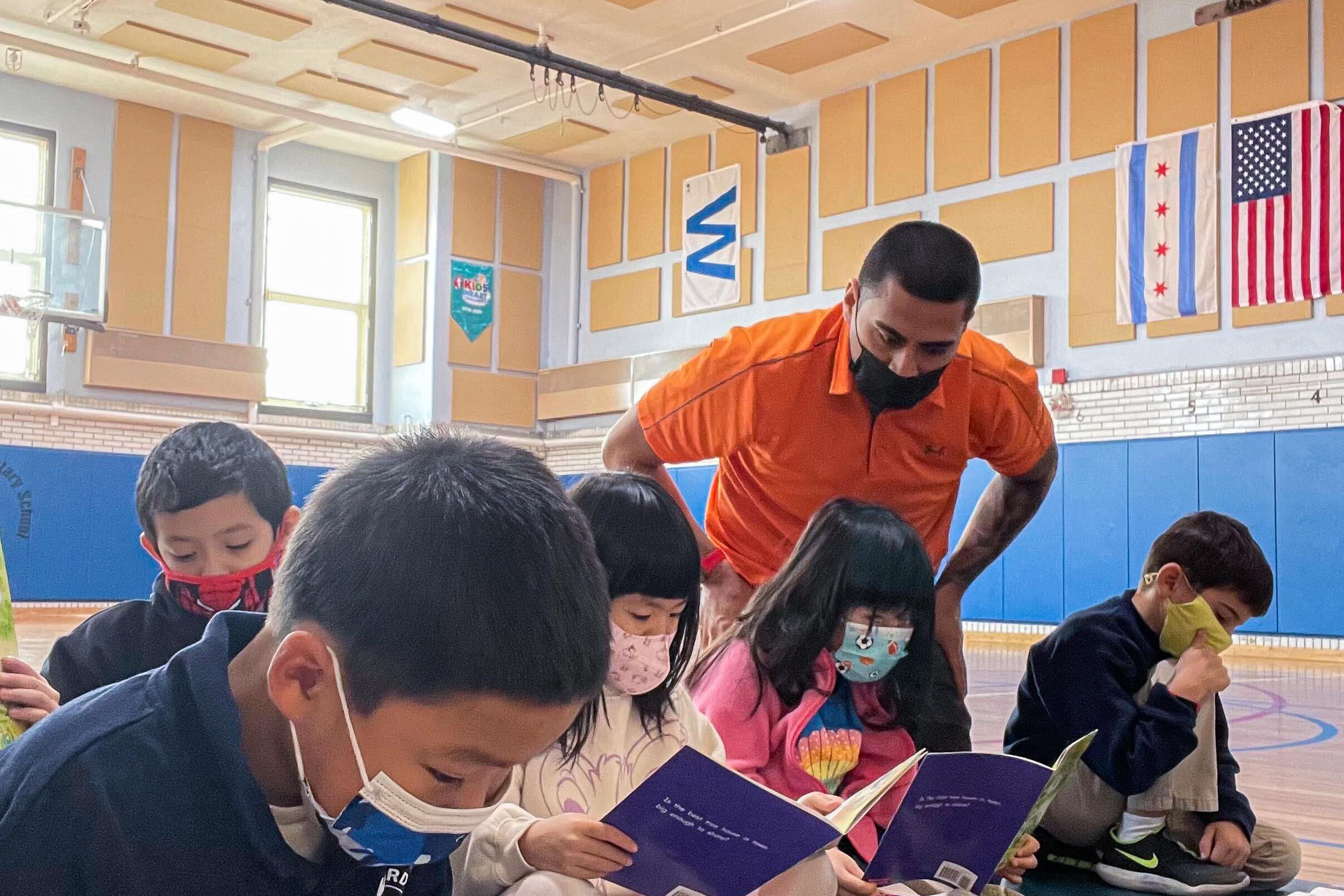 A teacher wearing an orange polo looks over the shoulders of several young students, who are reading in a school gymnasium and wearing protective masks.