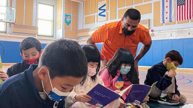A teacher wearing an orange polo looks over the shoulders of several young students, who are reading in a school gymnasium and wearing protective masks.
