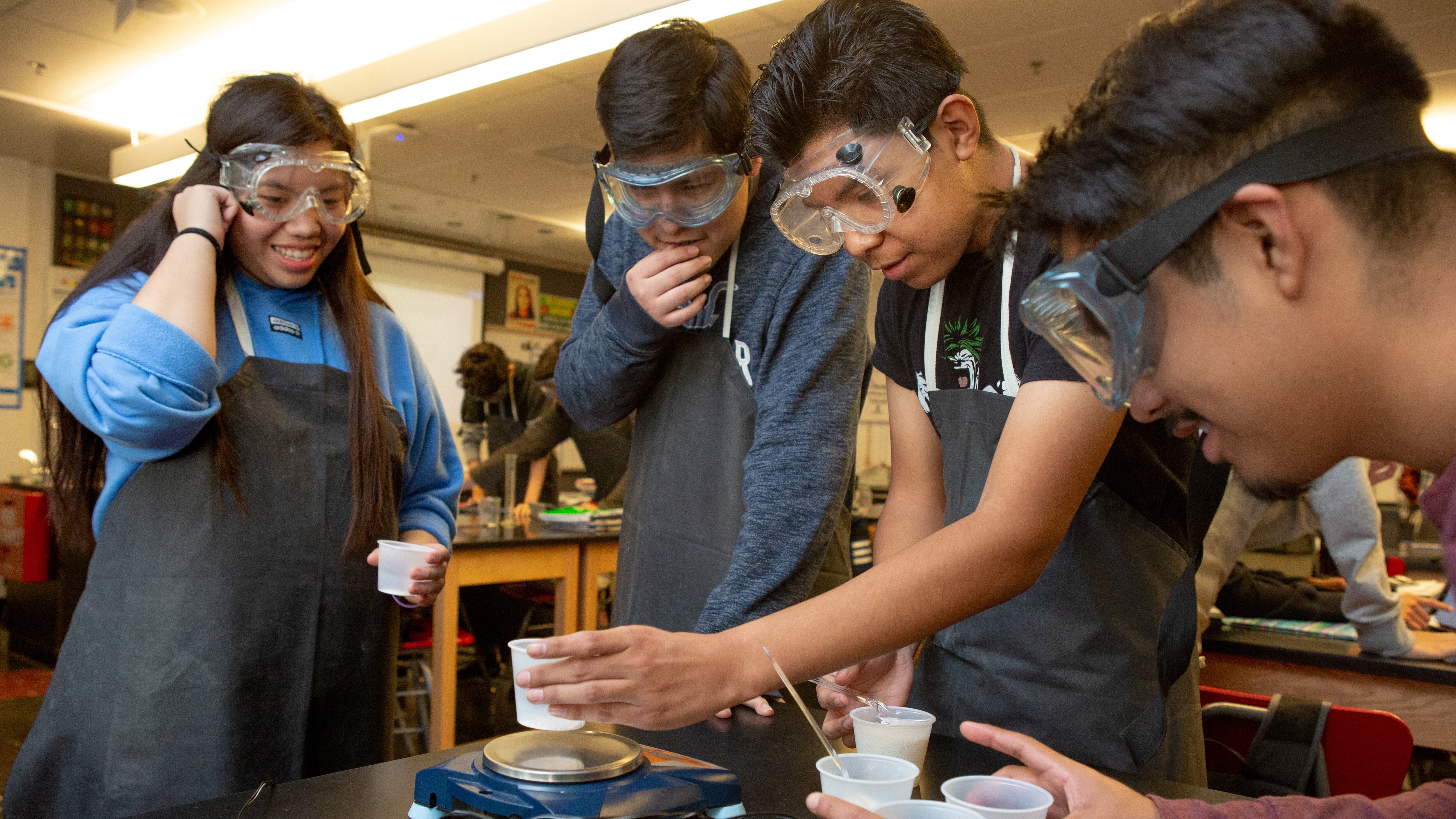 Four high school students wearing protective lab equipment work together on an experiment.
