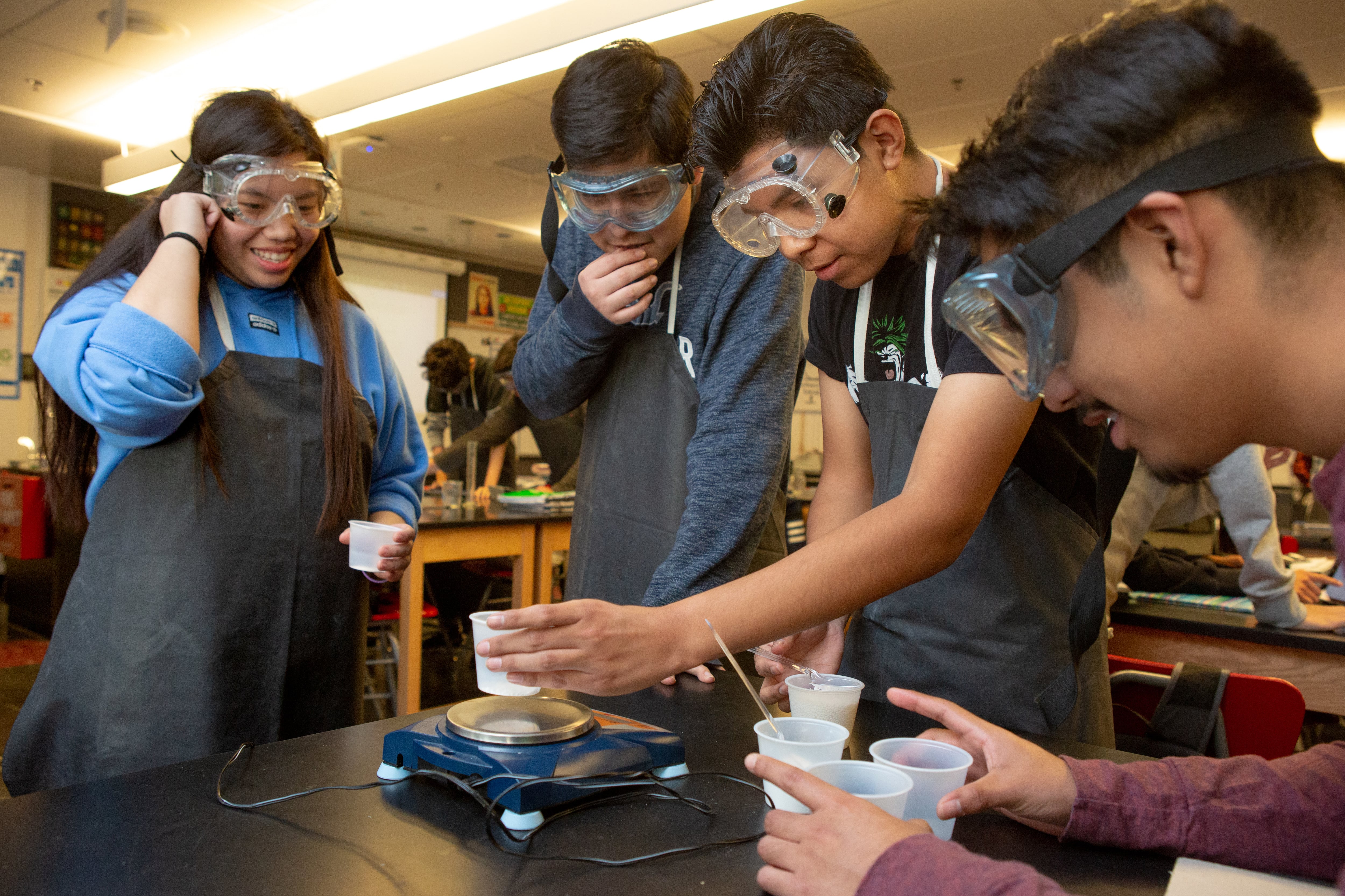 Four high school students wearing protective lab equipment work together on an experiment.