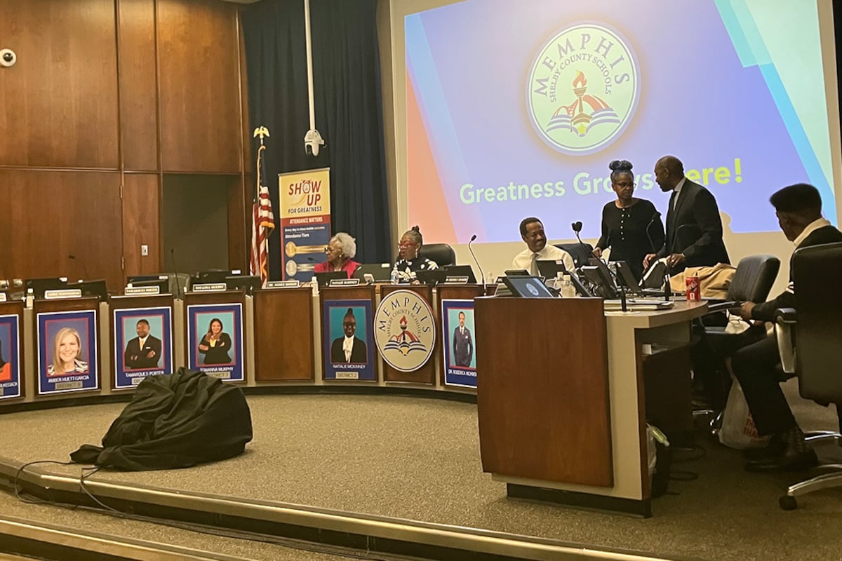 A photograph of a school board meeting with people in suits sitting and standing by a u-shape row of wooden desks.