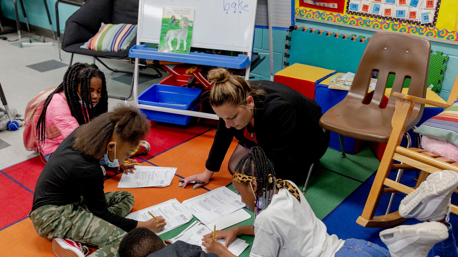 Four young students lay on a colorful rug as their teacher guides them through a worksheet.