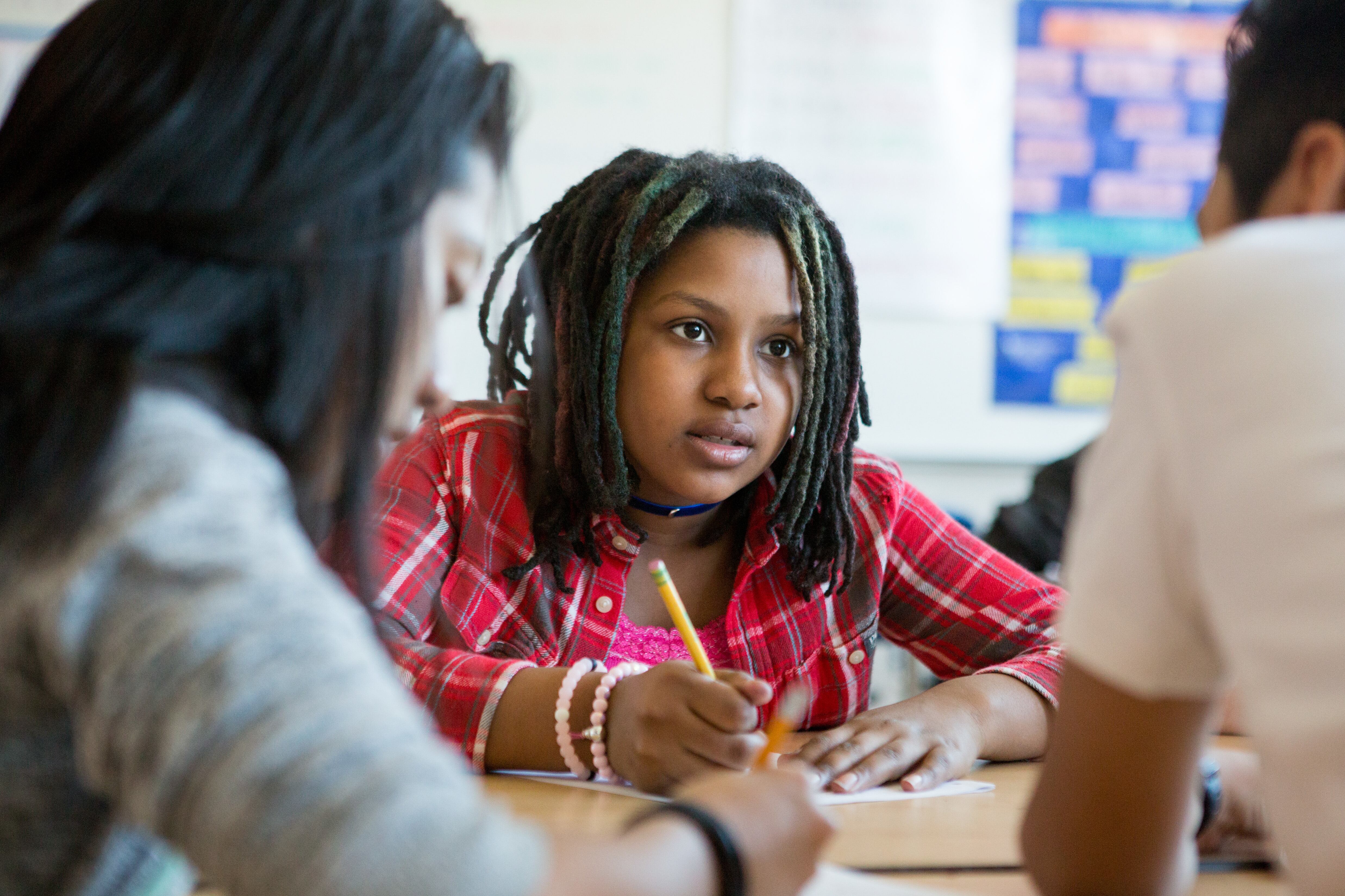 A young teenage girl with dreadlocks appears to be asking a question as she works on an assignment at a table with other students. We see her face clearly, while the other students have their backs to us and are slightly blurry.