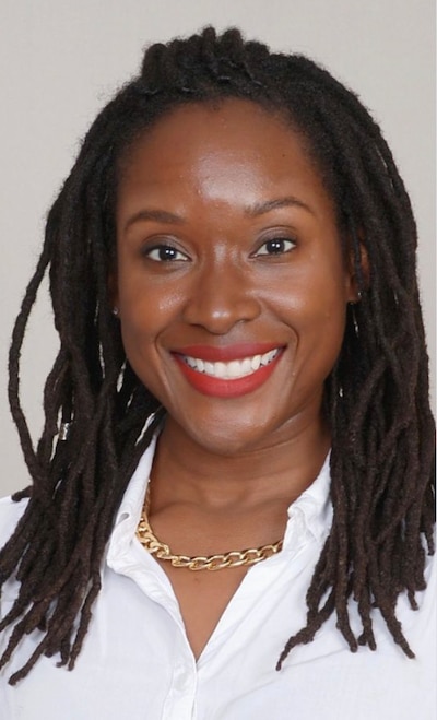 A photograph of a Black woman with long dark hair and wearing a white dress shirt and posing for a photograph while smiling.