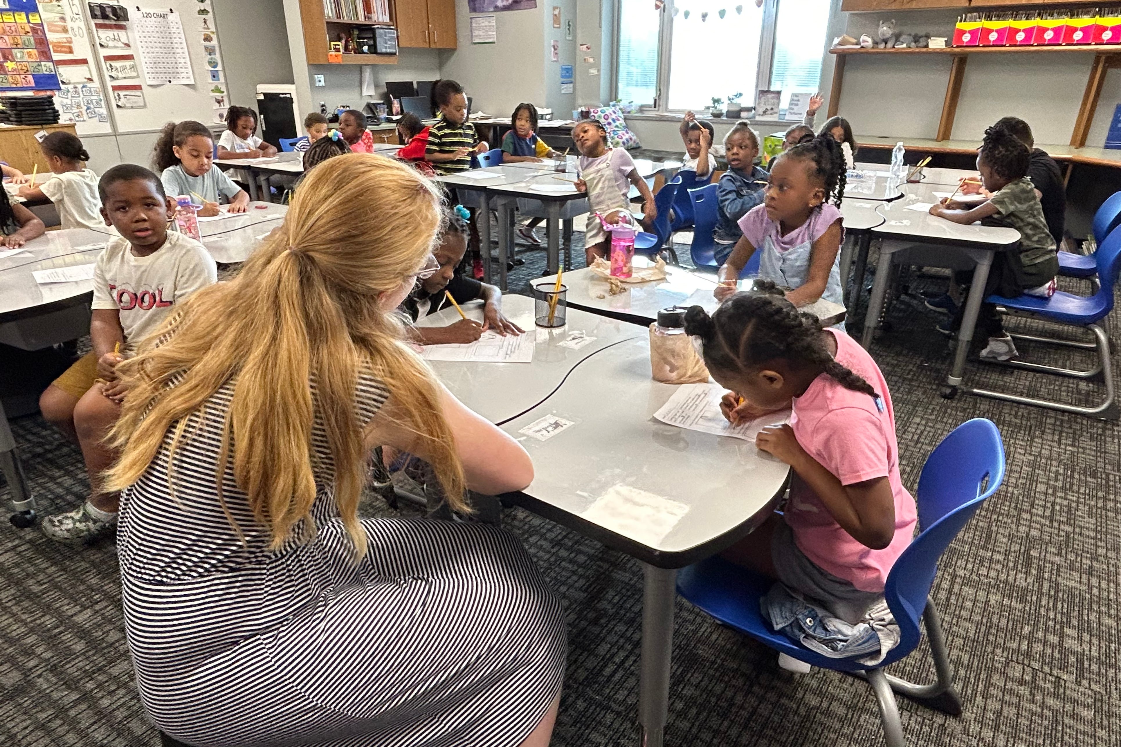 An adult with long blonde hair sits at a desk full of students in a room with desks full of students.