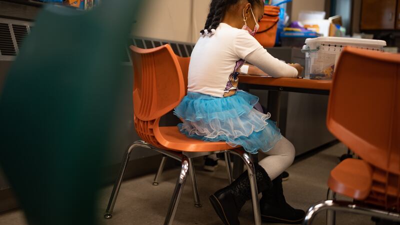 A young girl in a blue tutu skirt works at a desk, seen behind green and orange chairs.