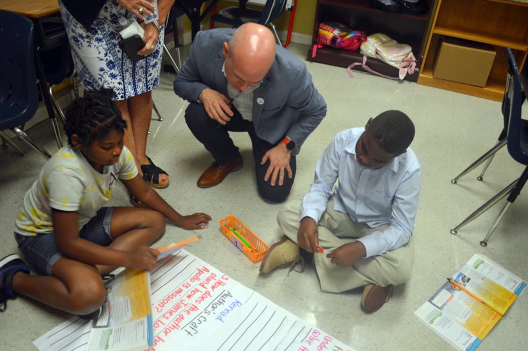 Two young students write on a poster board on the ground as an adult peers overhead to review their work.