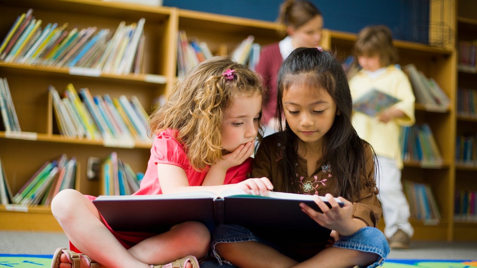Two little girls sit on the floor of a library read a book together.