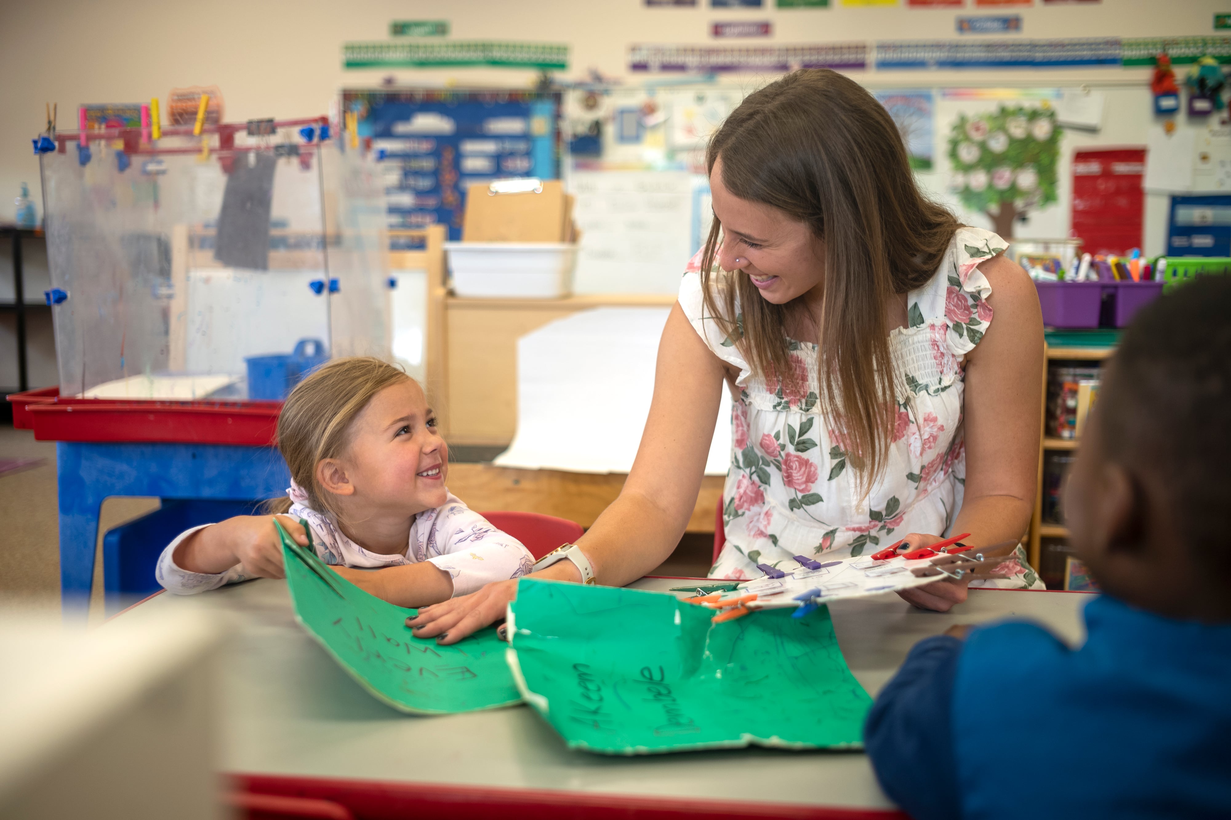 A preschool teacher in a white flowery dress looks at a little girl sitting next to her who is also smiling.
