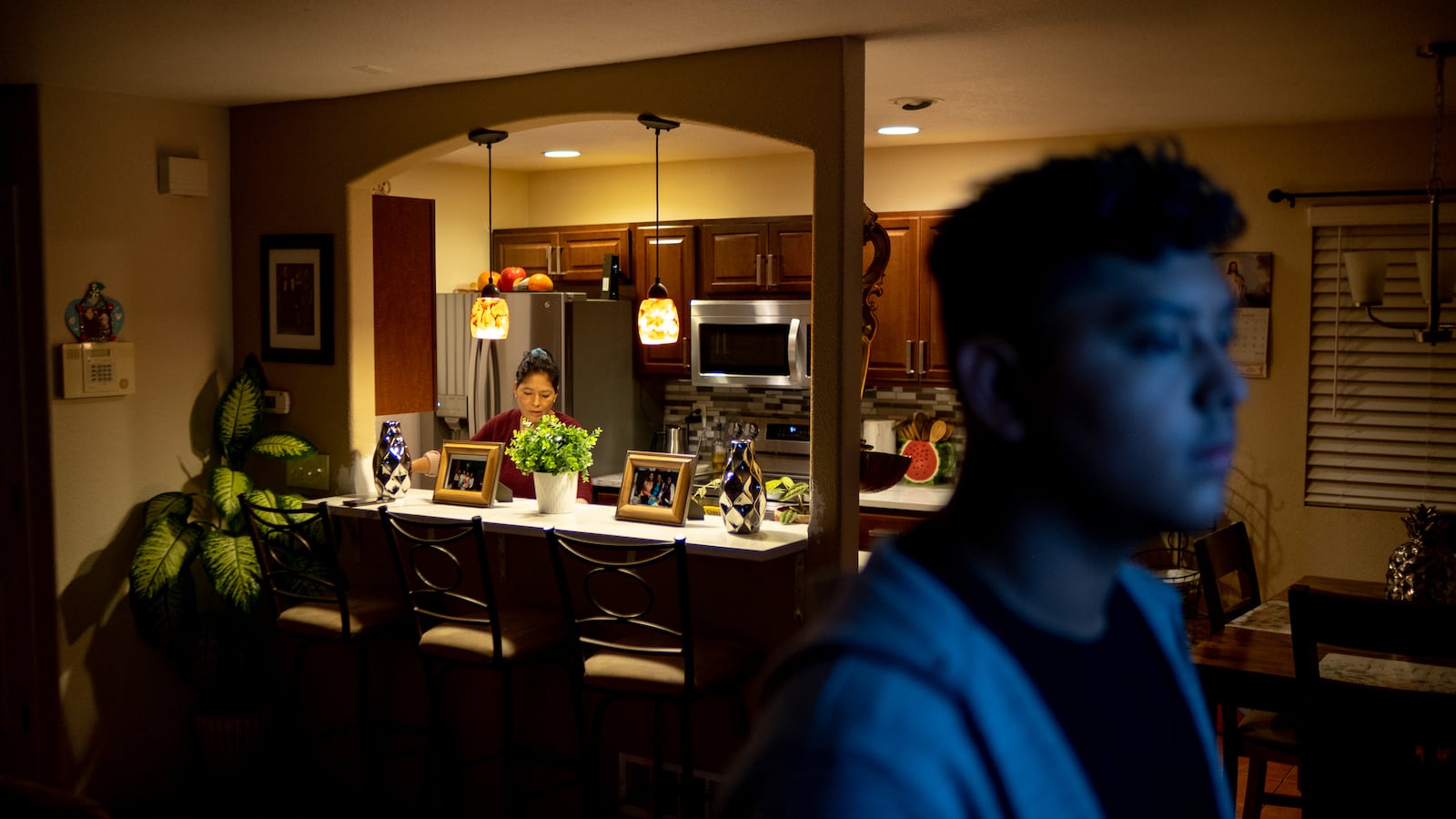 A mother works in the kitchen during an early morning as her son looks at the television in the living room, its blue light reflecting off of his face.