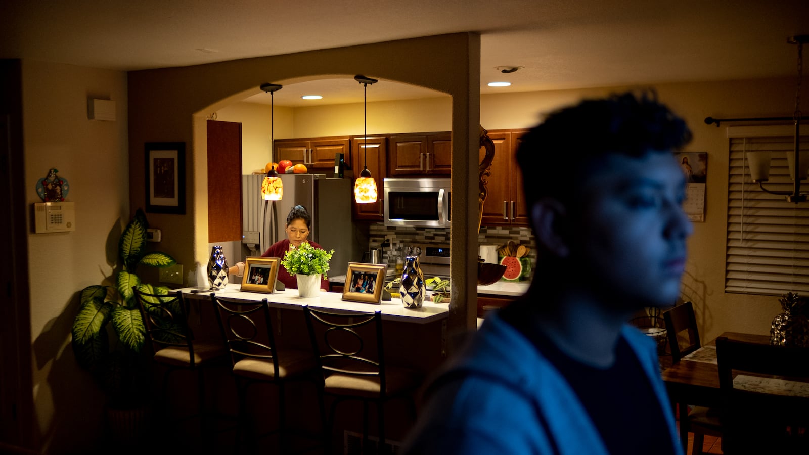 A mother works in the kitchen during an early morning as her son looks at the television in the living room, its blue light reflecting off of his face.
