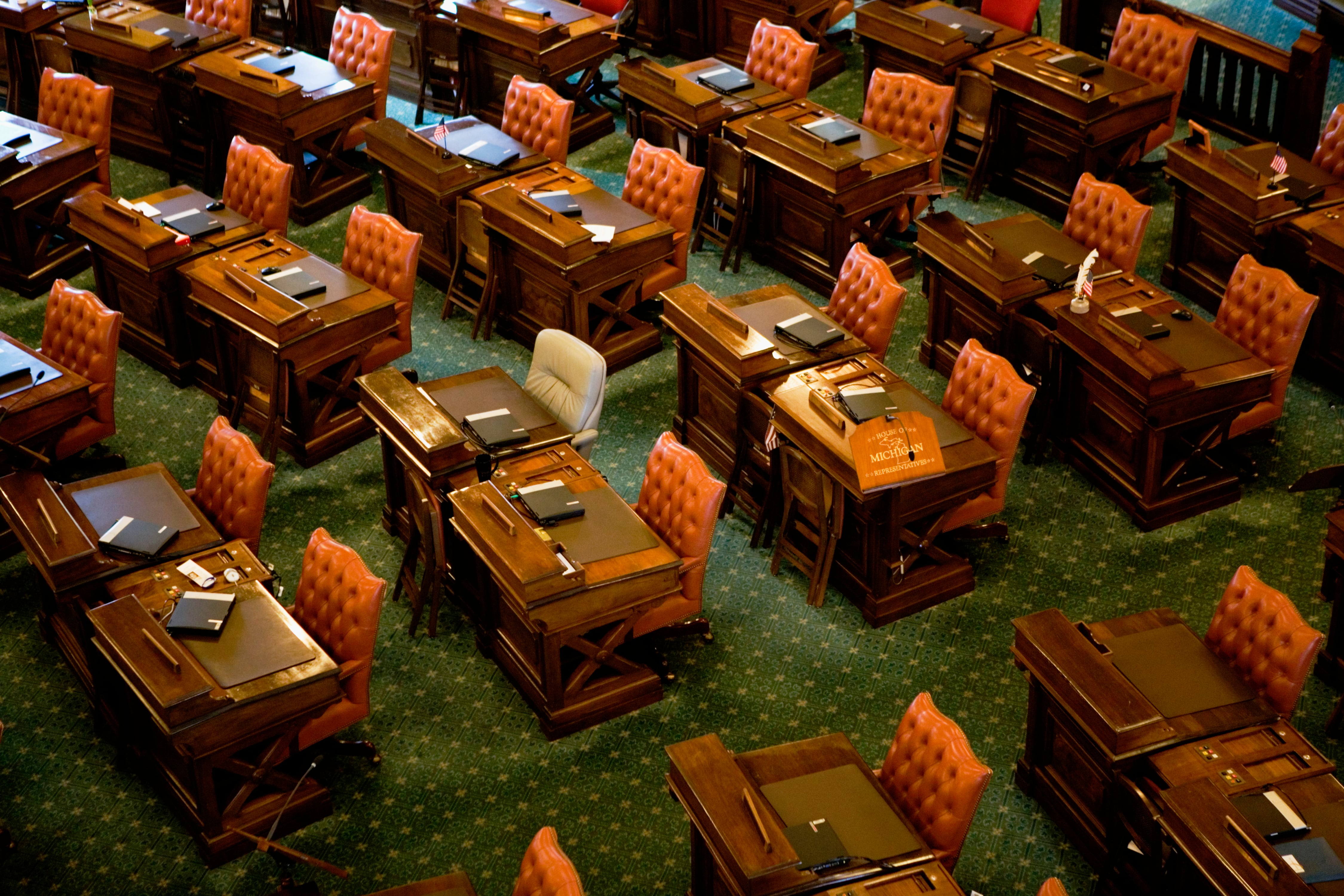 Five rows of wooden desks with orange chairs in a large room with green carpet.