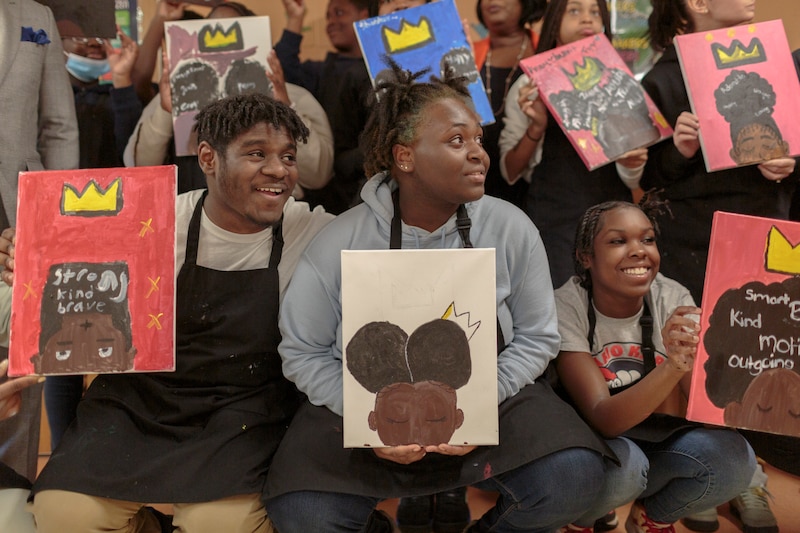 Several students wearing black smocks smile and hold up artwork featuring yellow crowns.