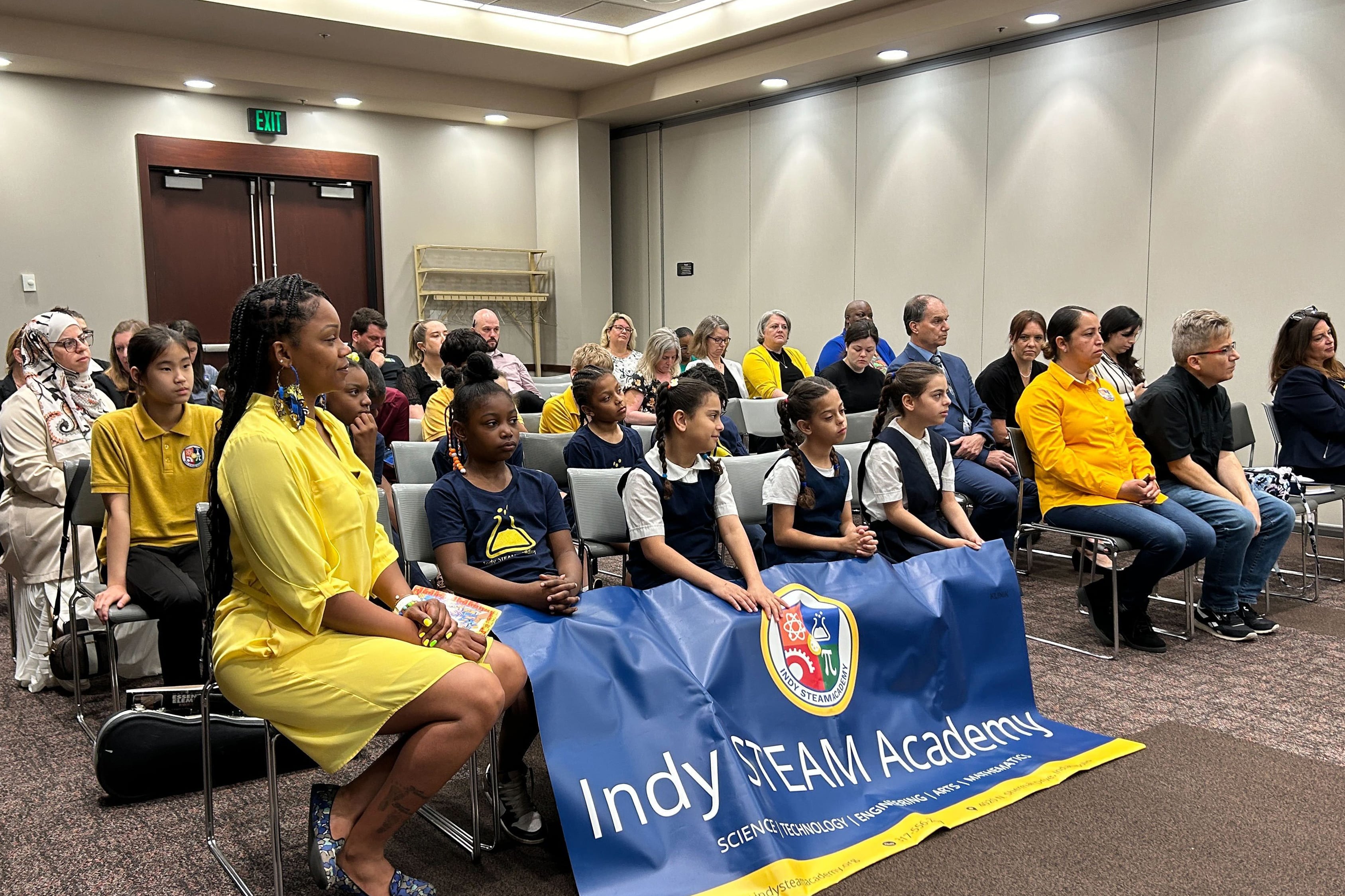 A group of young students sit in the front row with an adult while the chairs in the background are full of people. They are in what looks like a conference room.