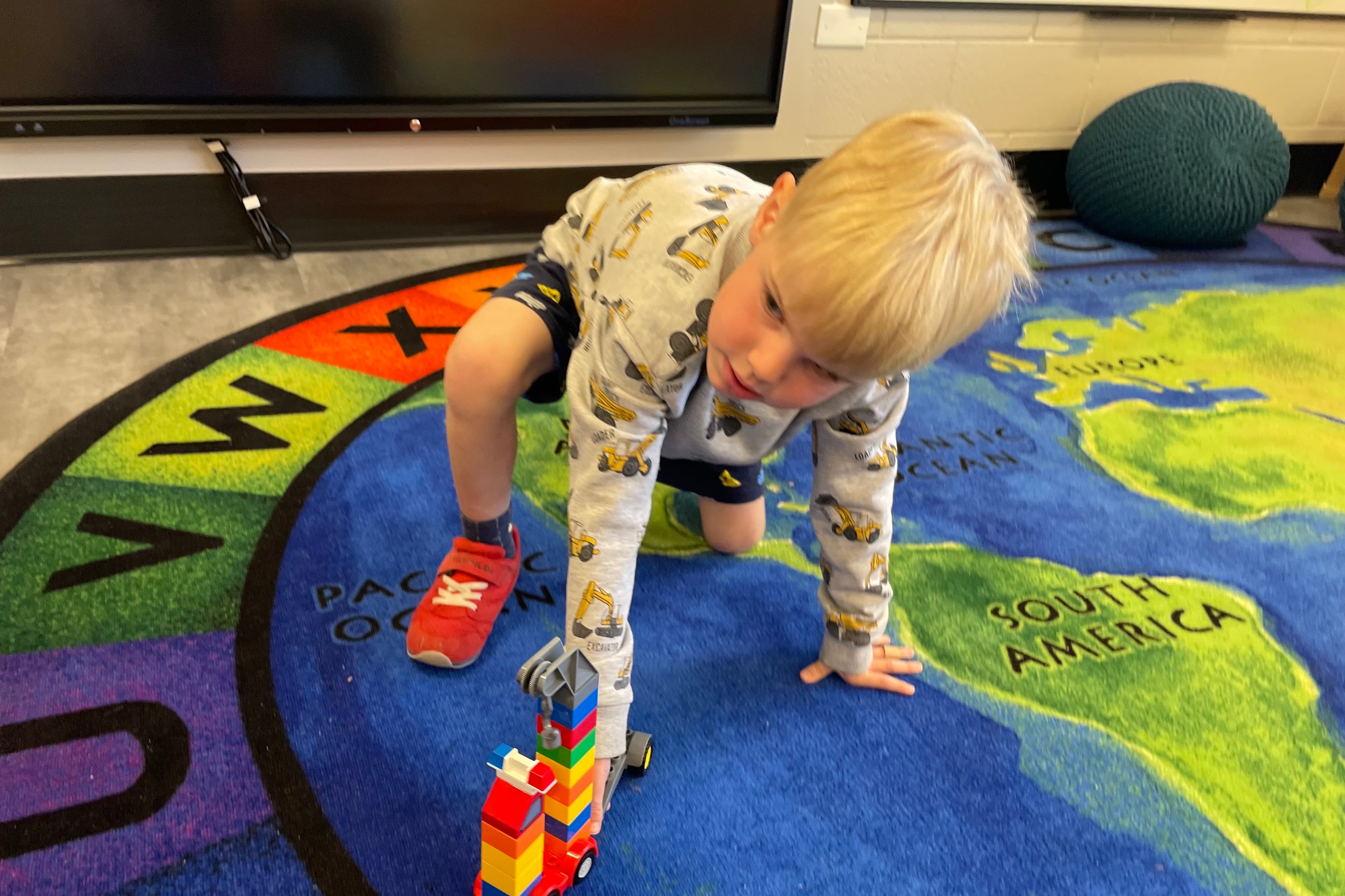A young boy kneels on a colorful carpet and rolls a Lego truck.