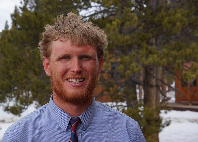A photograph of a white man with a beard and wearing a blue shirt and tie standing for a portrait in front of green trees.