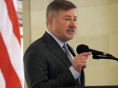 A man wearing a suit stands next to an American flag and speaks into a microphone.