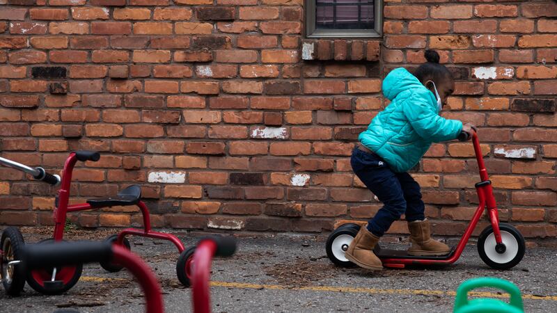 A little girl rides around on a scooter in front of a red brick wall, past other scooters and tricycles in the foreground.