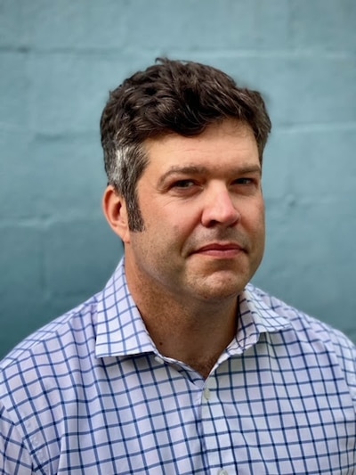 A man in a plaid dress shirt with short brown hair poses for a portrait in front of a blue brick wall.