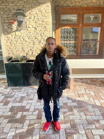 A young person wearing a black coat stands in front of a door holding a coffee mug.