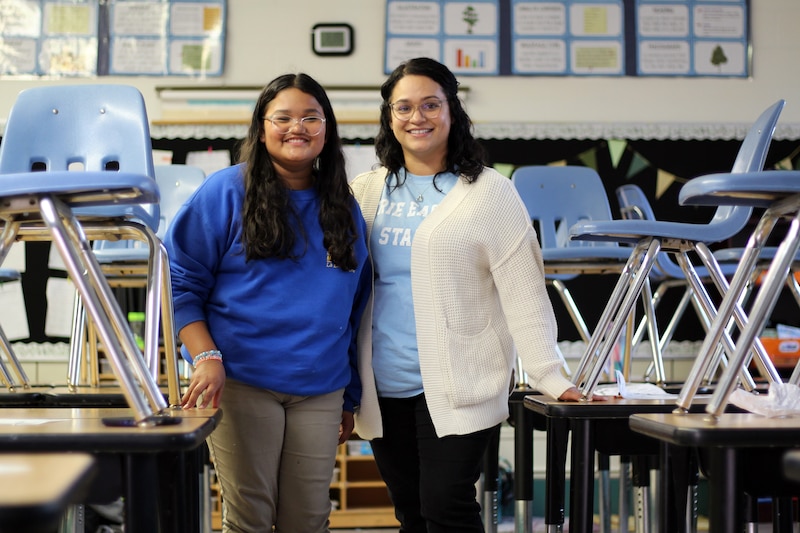 A middle school girl and an adult, both with dark hair stand side by side in a classroom with chairs stacked on the tables.