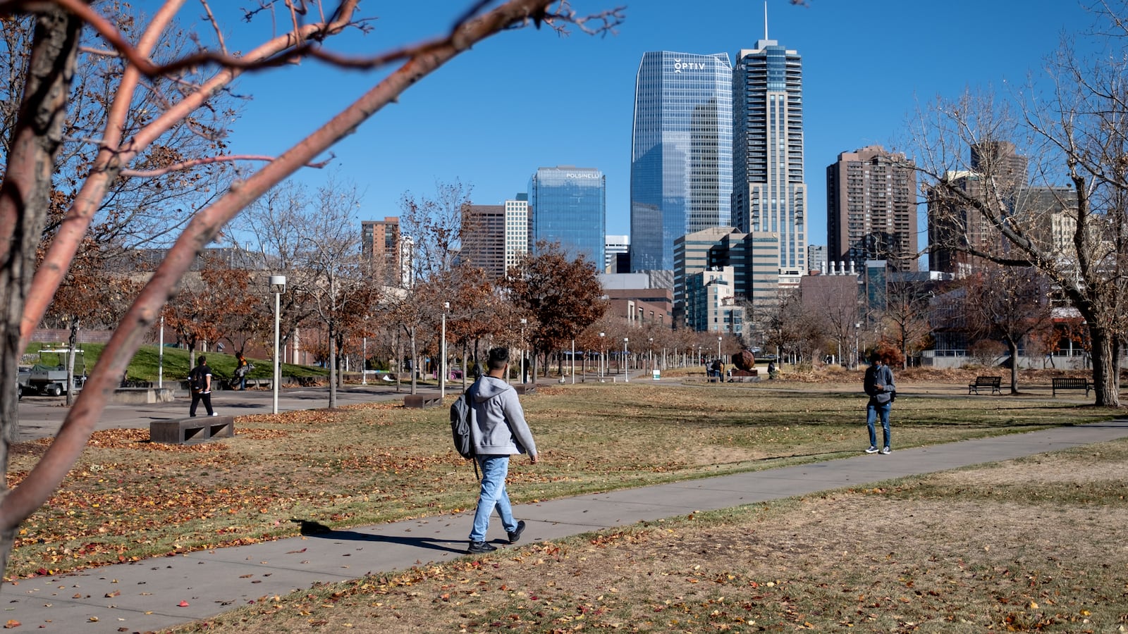 A young man, wearing a grey sweatshirt and blue jeans, walks down a path toward campus, the Denver skyline looming in the background.