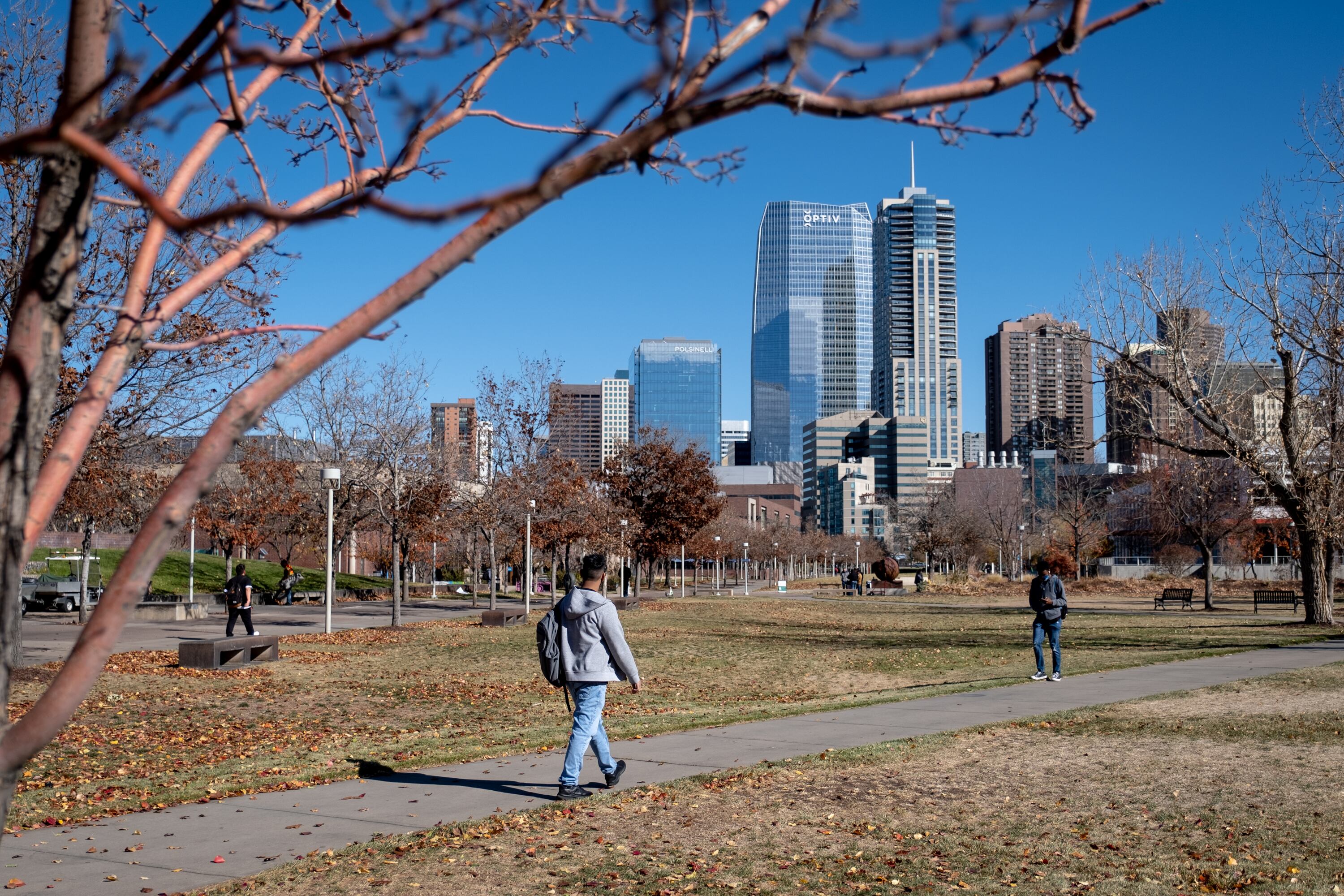 A young man, wearing a grey sweatshirt and blue jeans, walks down a path toward campus, the Denver skyline looming in the background.