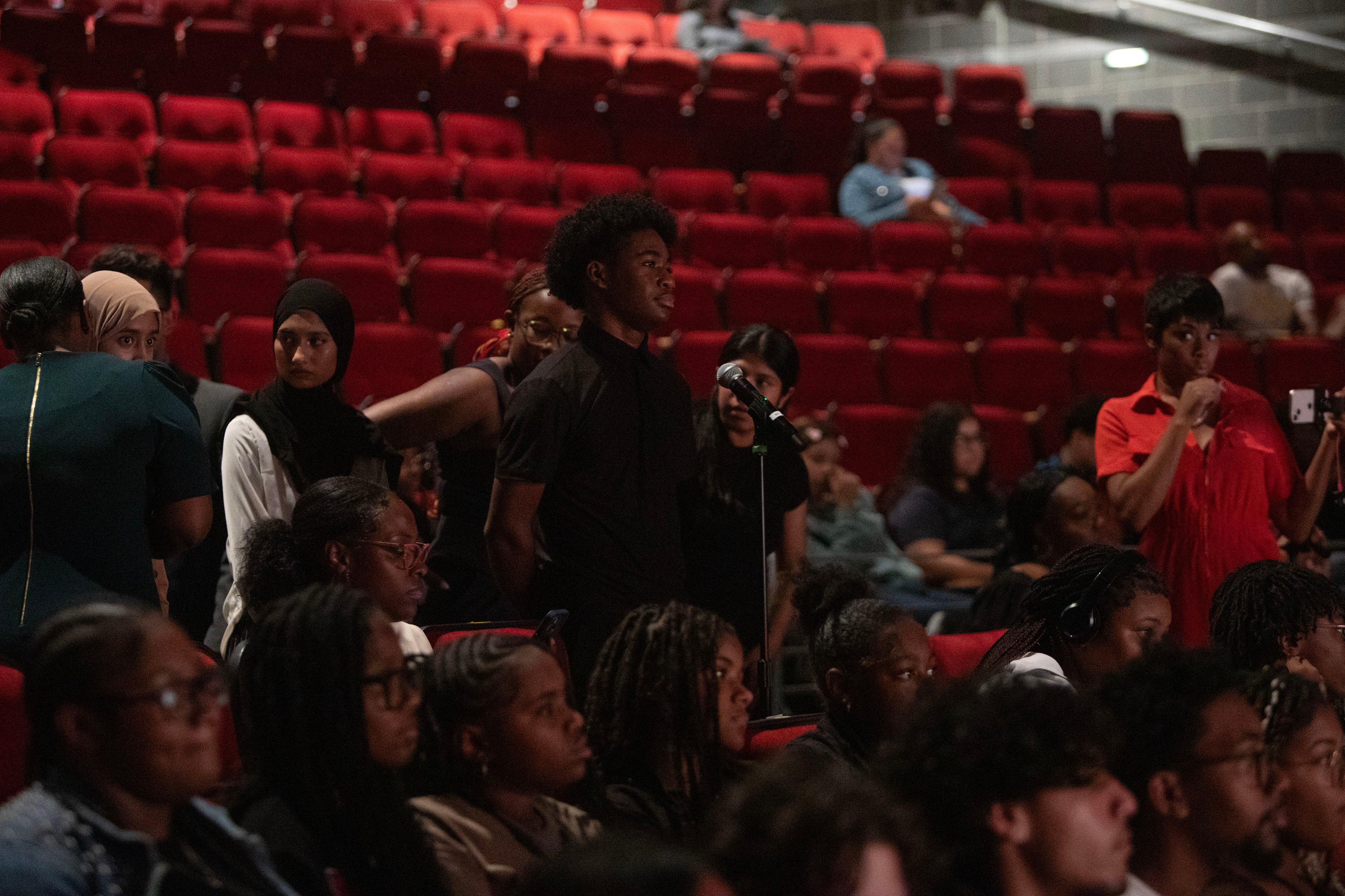 A high school student with short black hair stands behind a microphone and in front of a group of students in a dark auditorium.
