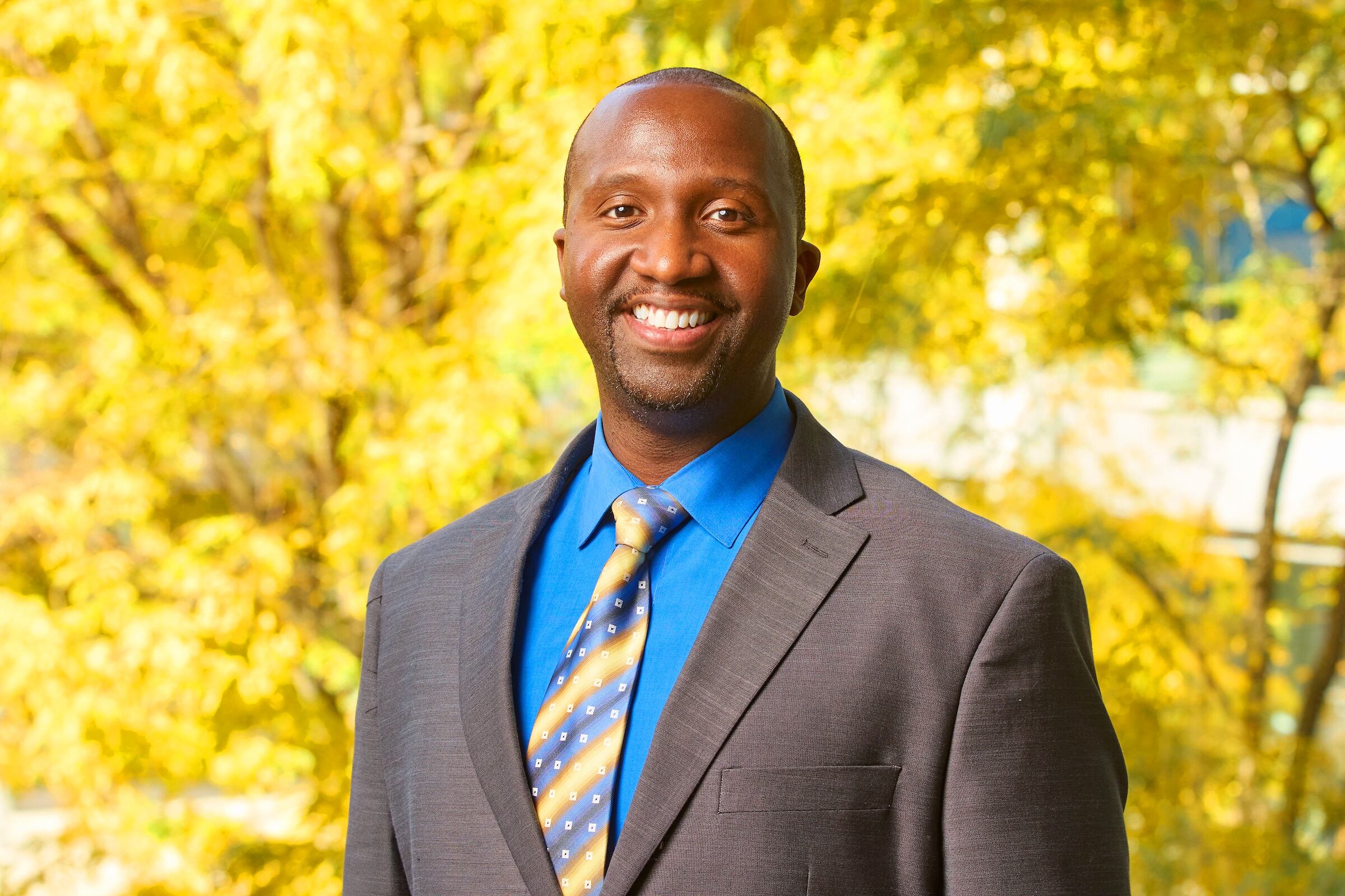 A man in a suit and tie stands smiling at the camera with trees in the background.