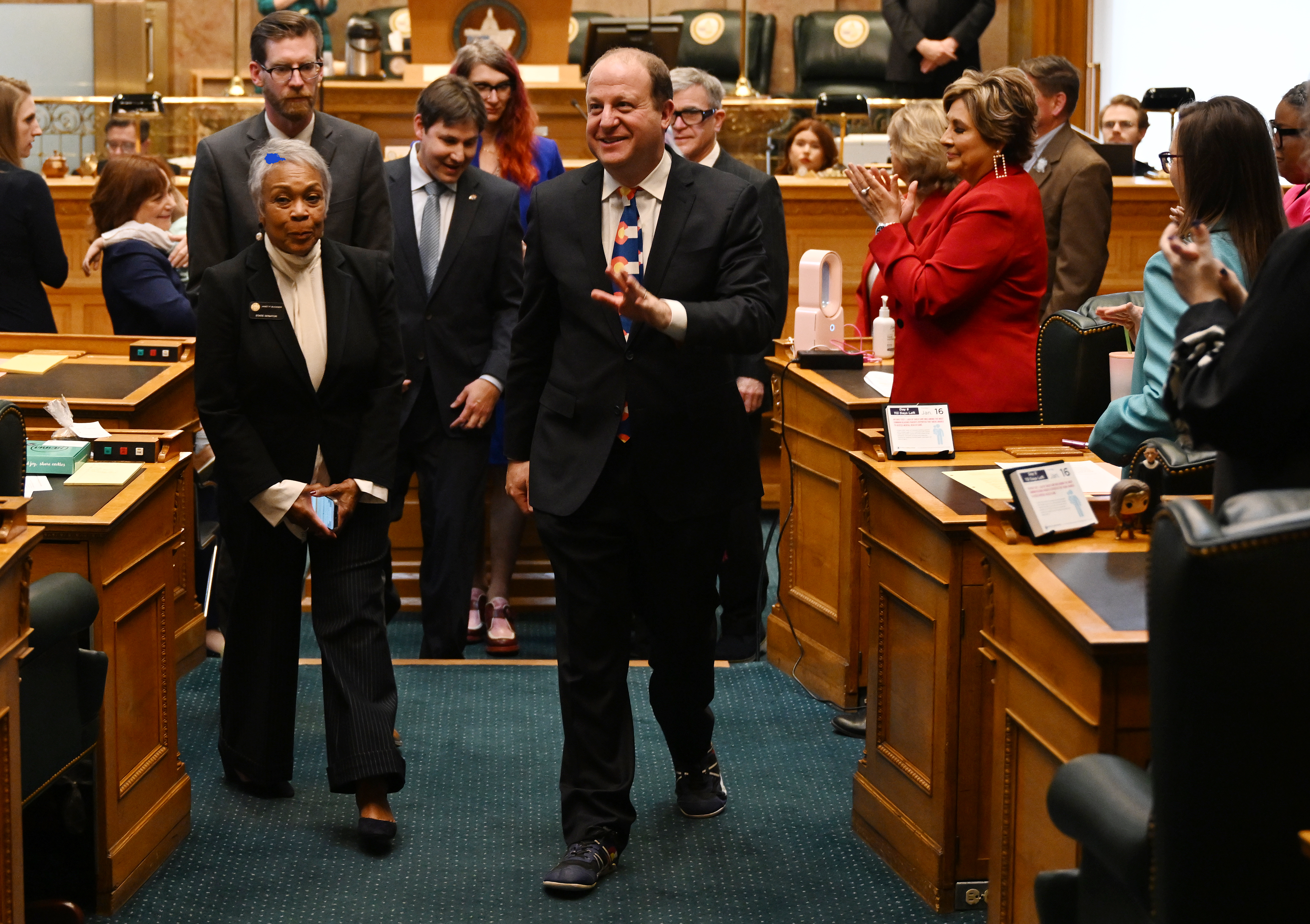 Colorado Gov. Jared Polis walks on a green carpet in the Colorado House chambers and surrounded by lawmakers while wearing a Colorado flag tie and black suit.