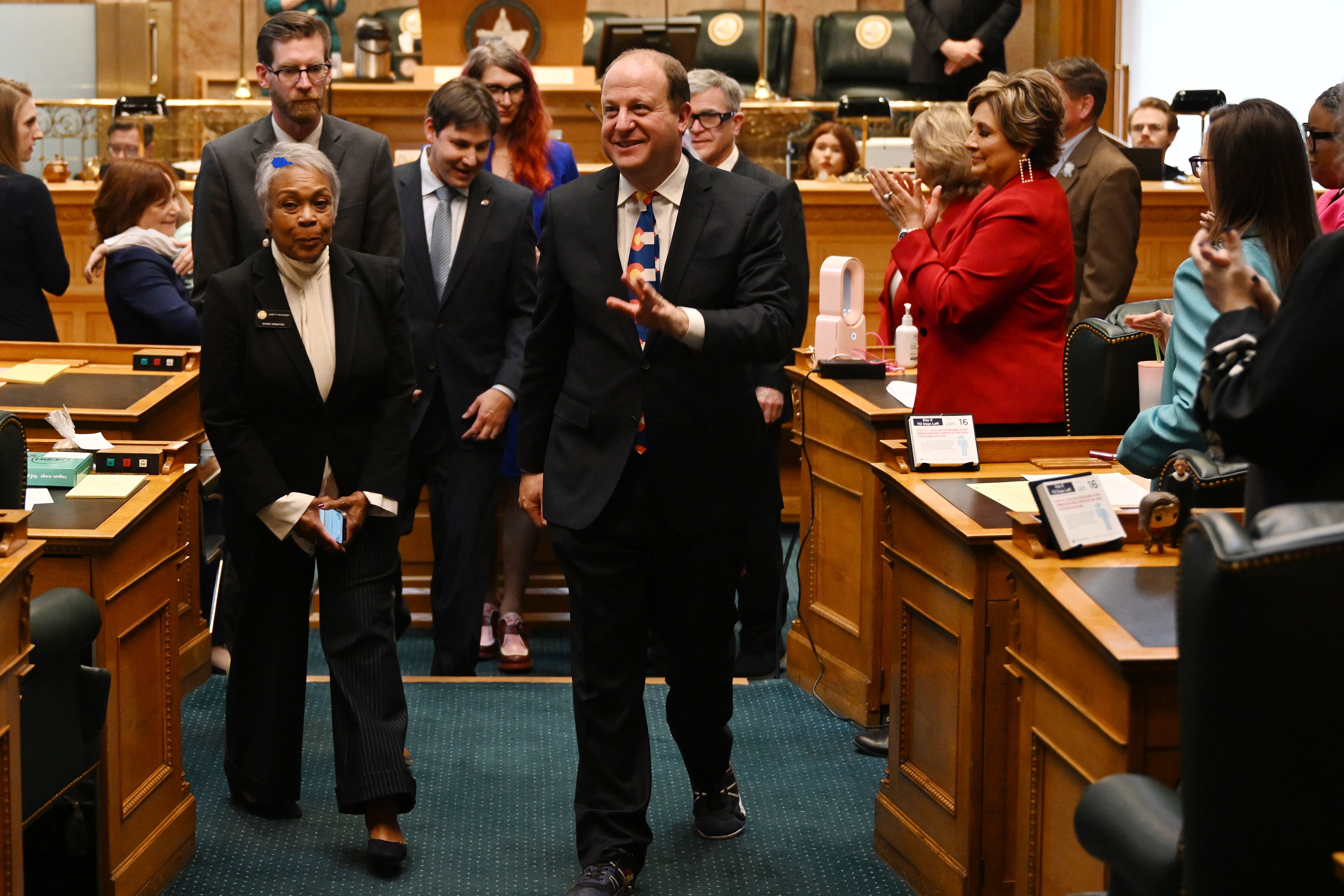 Colorado Gov. Jared Polis walks on a green carpet in the Colorado House chambers and surrounded by lawmakers while wearing a Colorado flag tie and black suit.