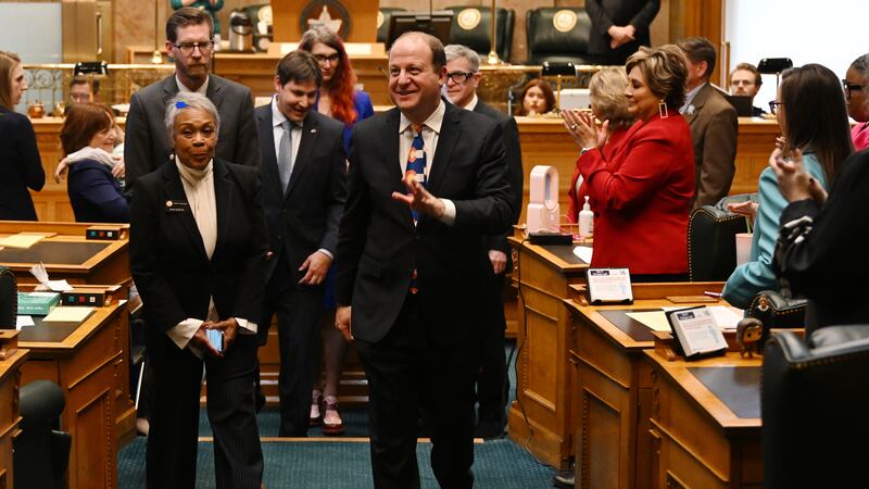 Colorado Gov. Jared Polis walks on a green carpet in the Colorado House chambers and surrounded by lawmakers while wearing a Colorado flag tie and black suit.