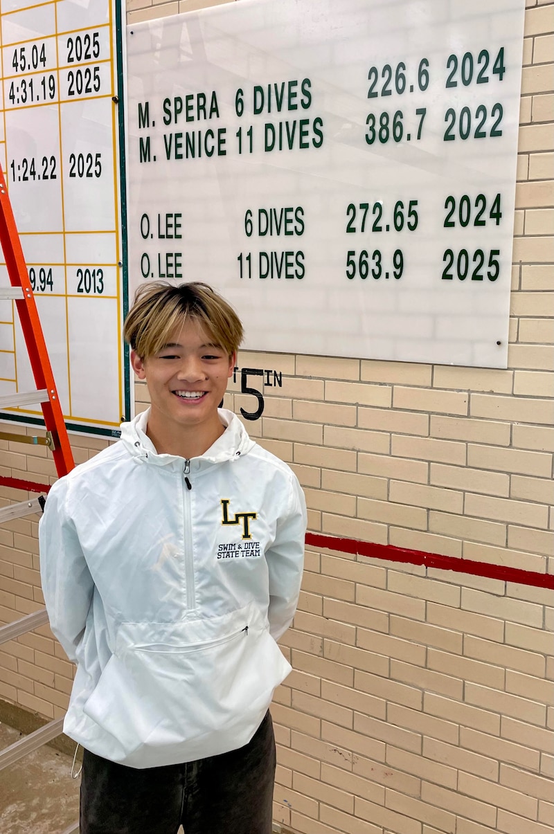A high school senior boy in a white jacket poses for a portrait in front of a white time board in a gym.
