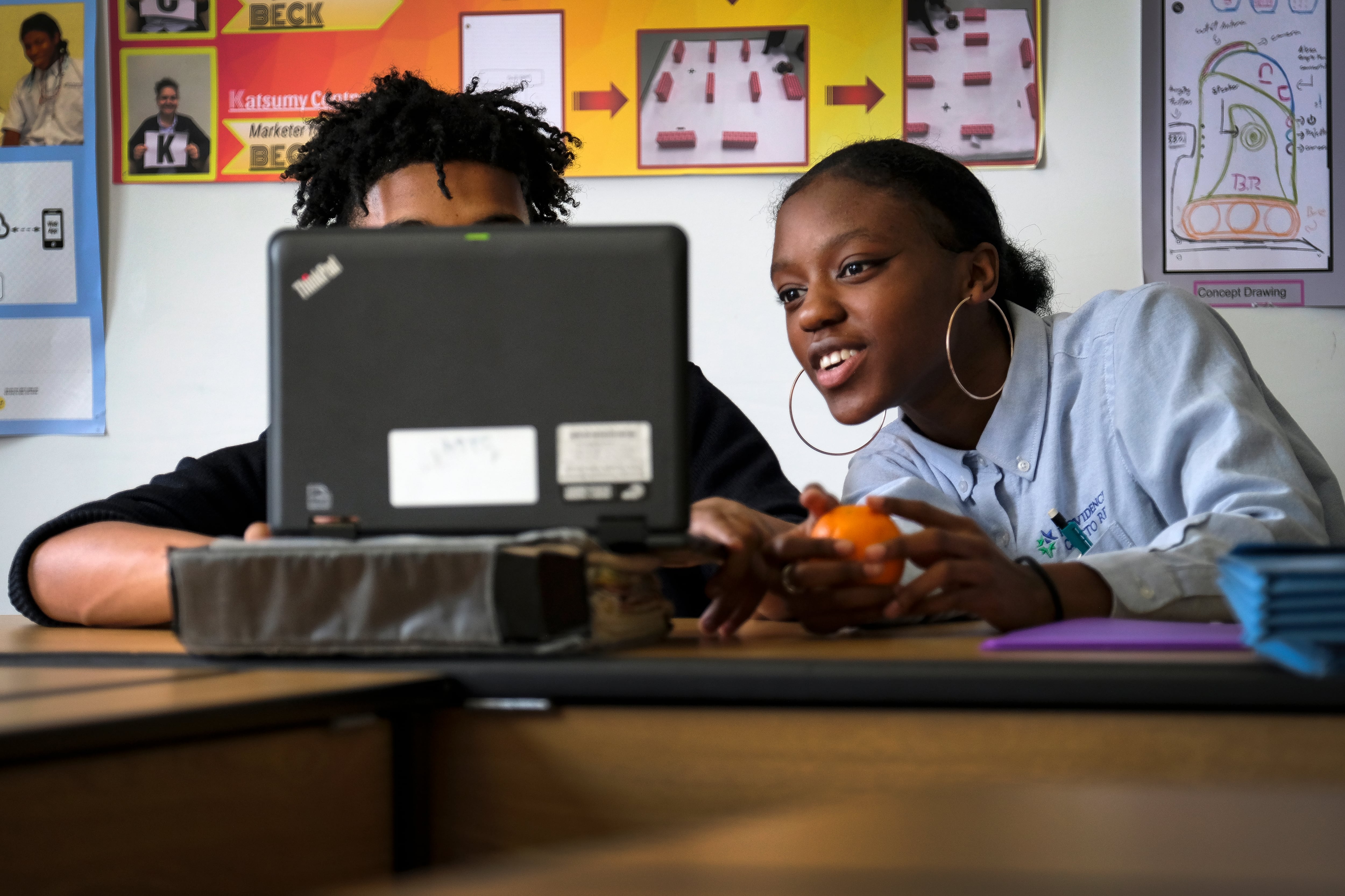 Two students sit at a wooden desk with school posters in the background. The student on the left is covered by a laptop and the student on the right is talking to the student and looking at the computer.