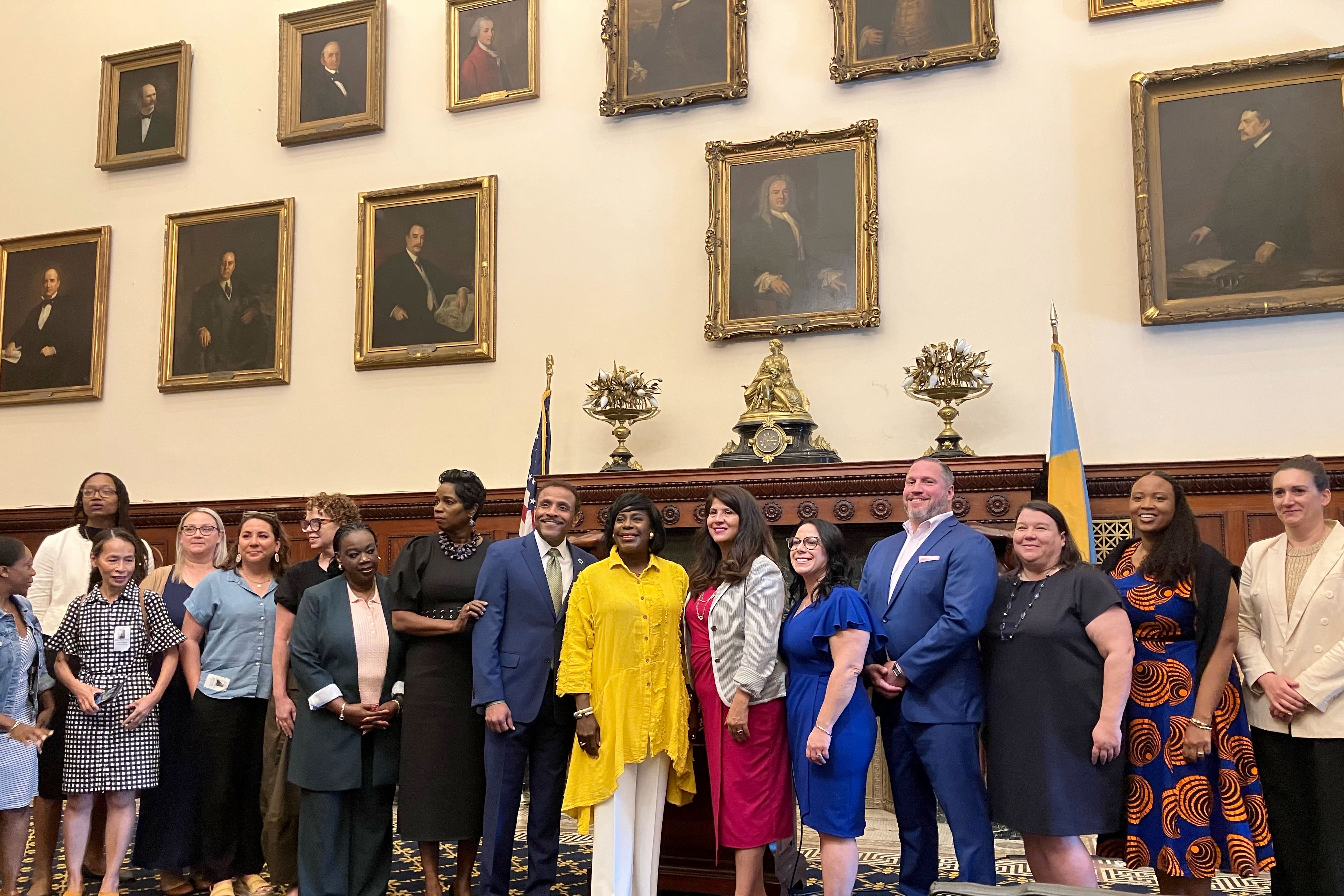A line of people stand in a line for a portrait in a large room with portraits of men on the wall in the background.