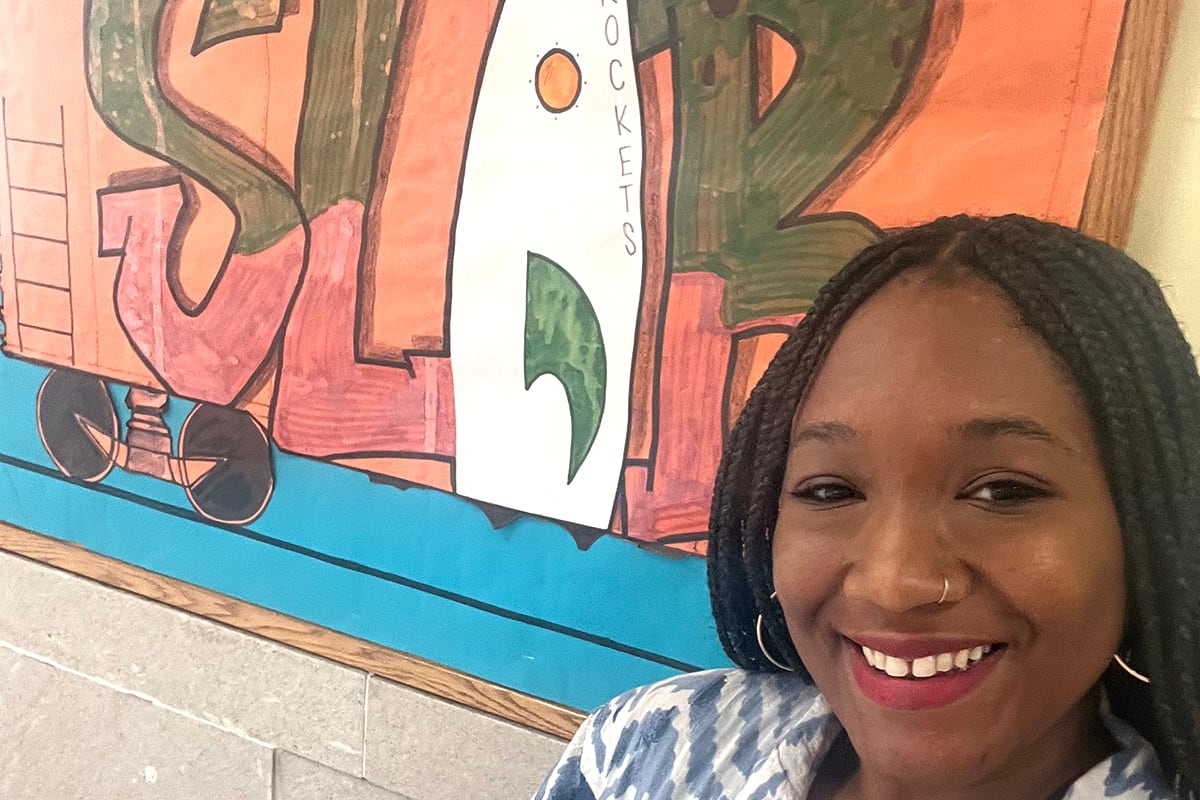 A woman in a blue and white top and dark hair stands in front of a bulletin board.