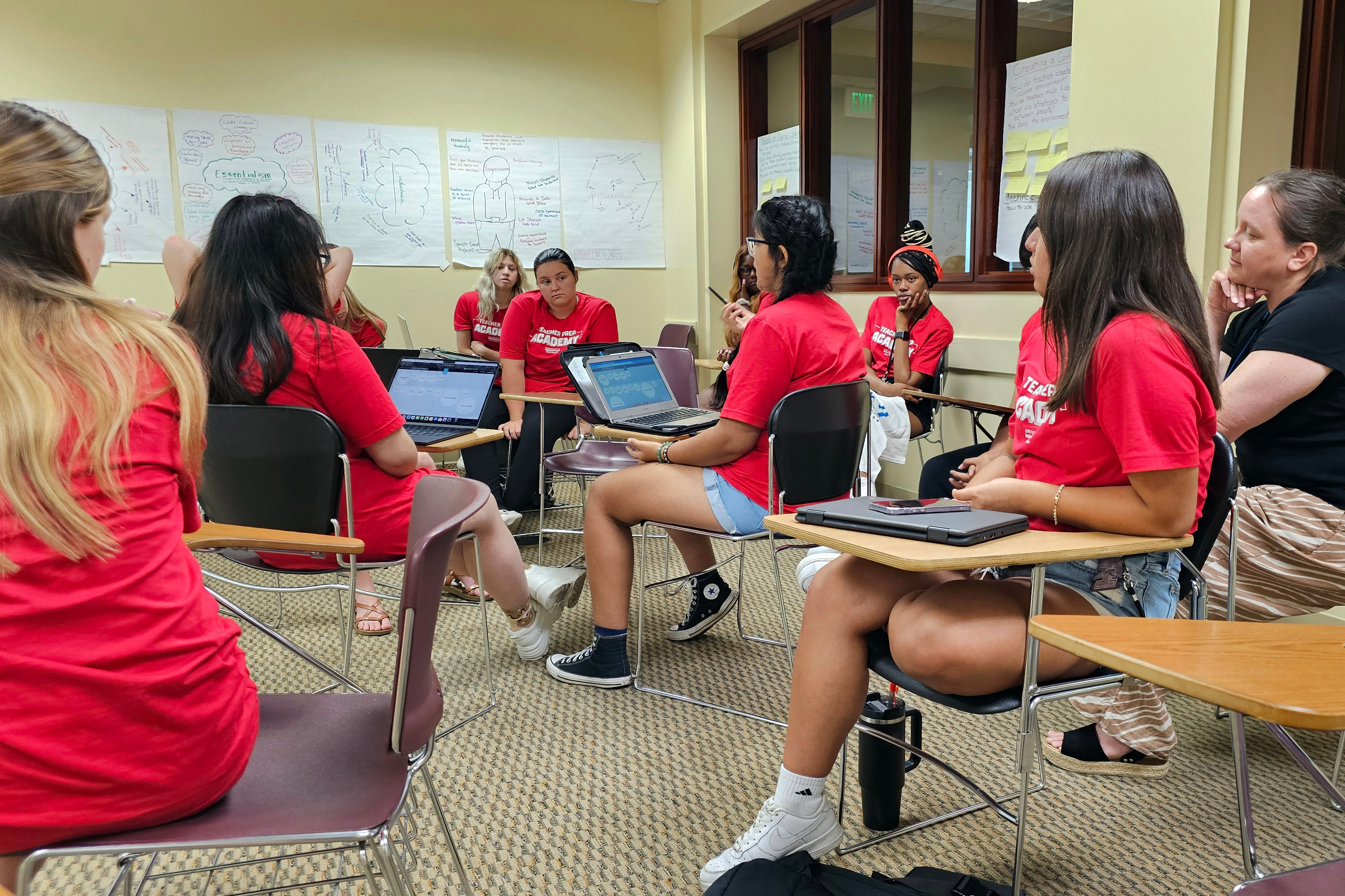 A person wearing a black shirt stands and speaks to a group of people sitting at desks wearing red t-shirts in a classroom.