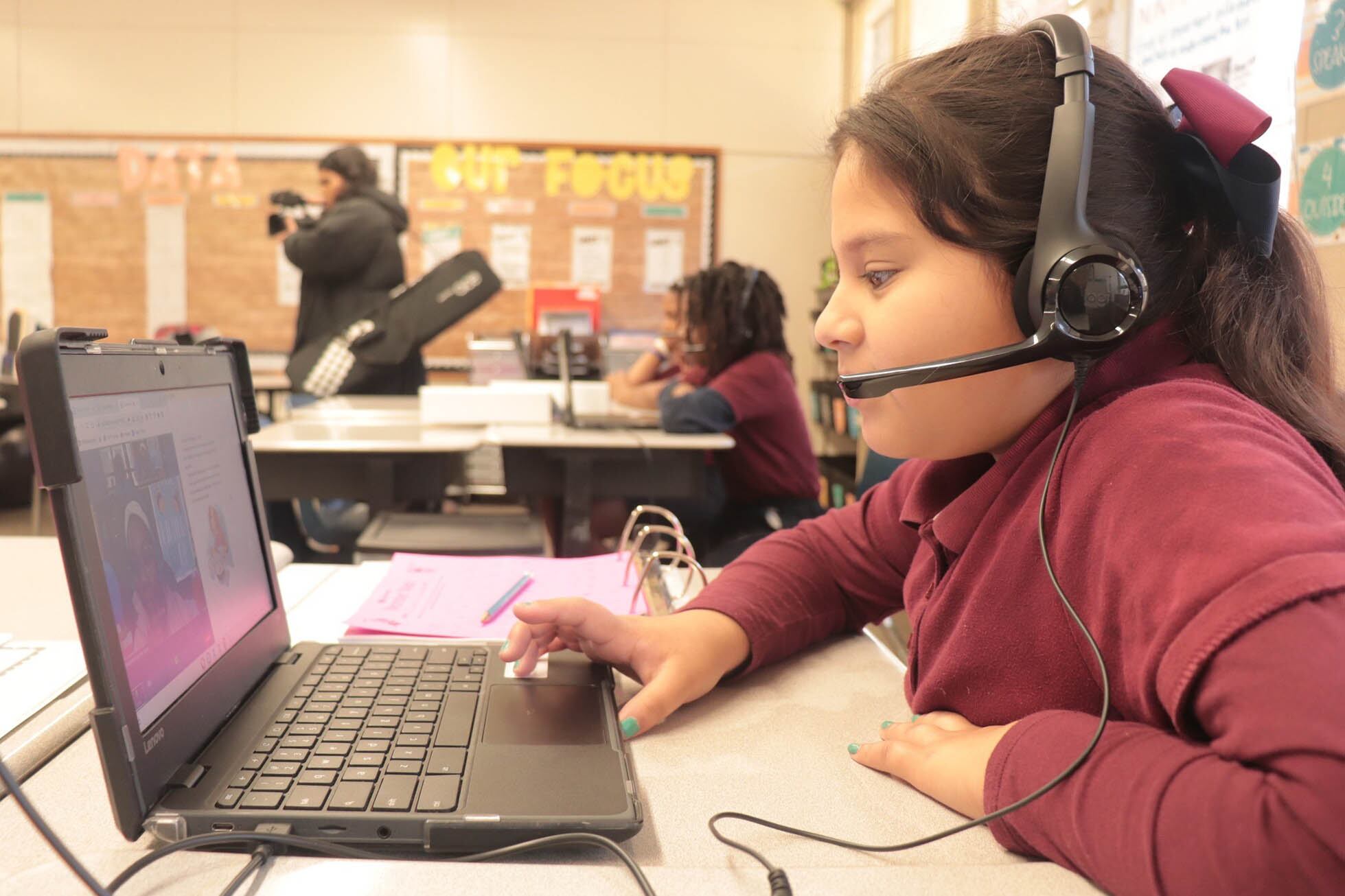 A young student with long dark hair and wearing a red shirt and headphones sits at a desk working on a laptop while an adult and other students are in the background.