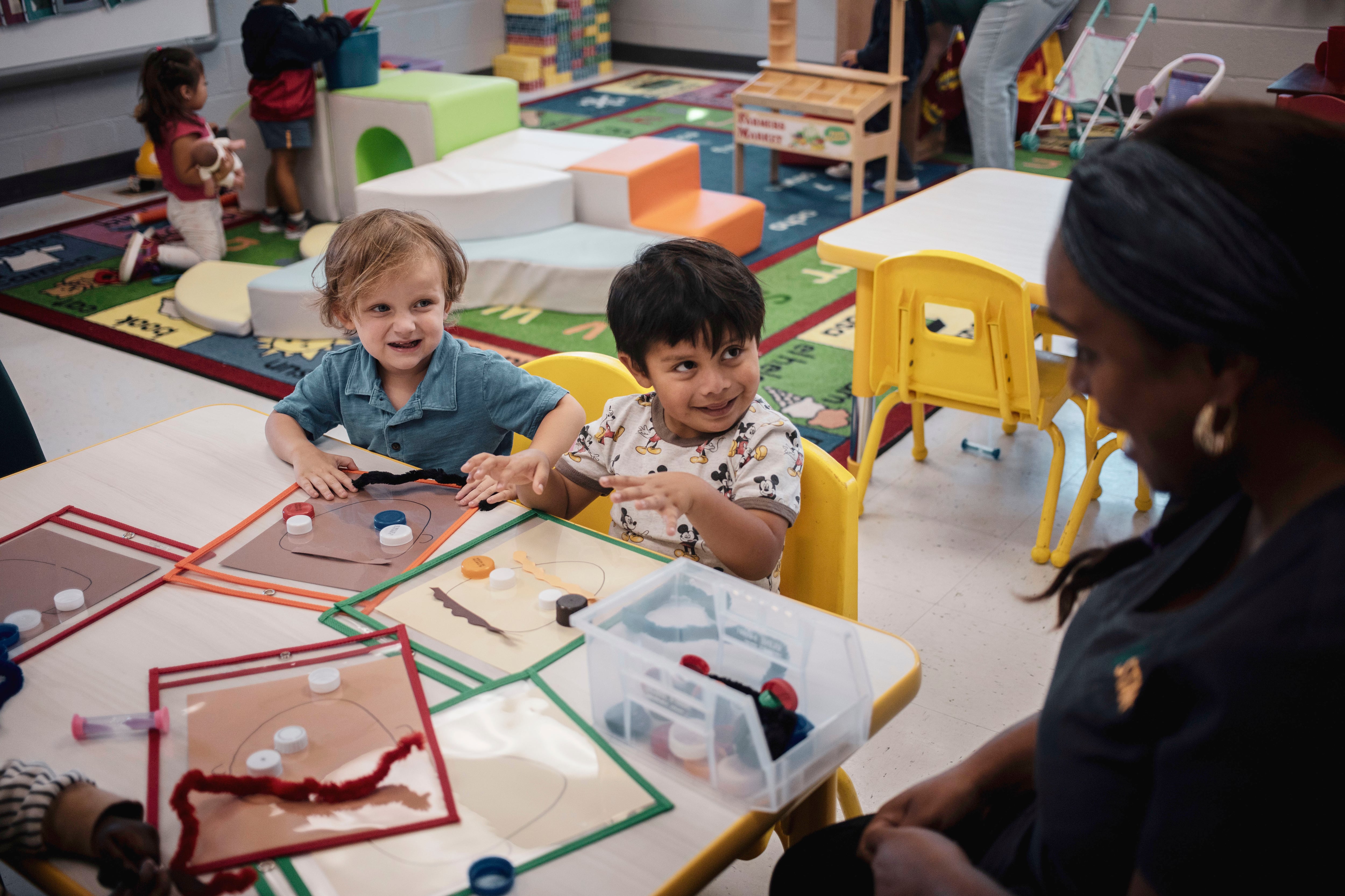 Two young boys smile up at a teacher sitting at table with them in a classroom.
