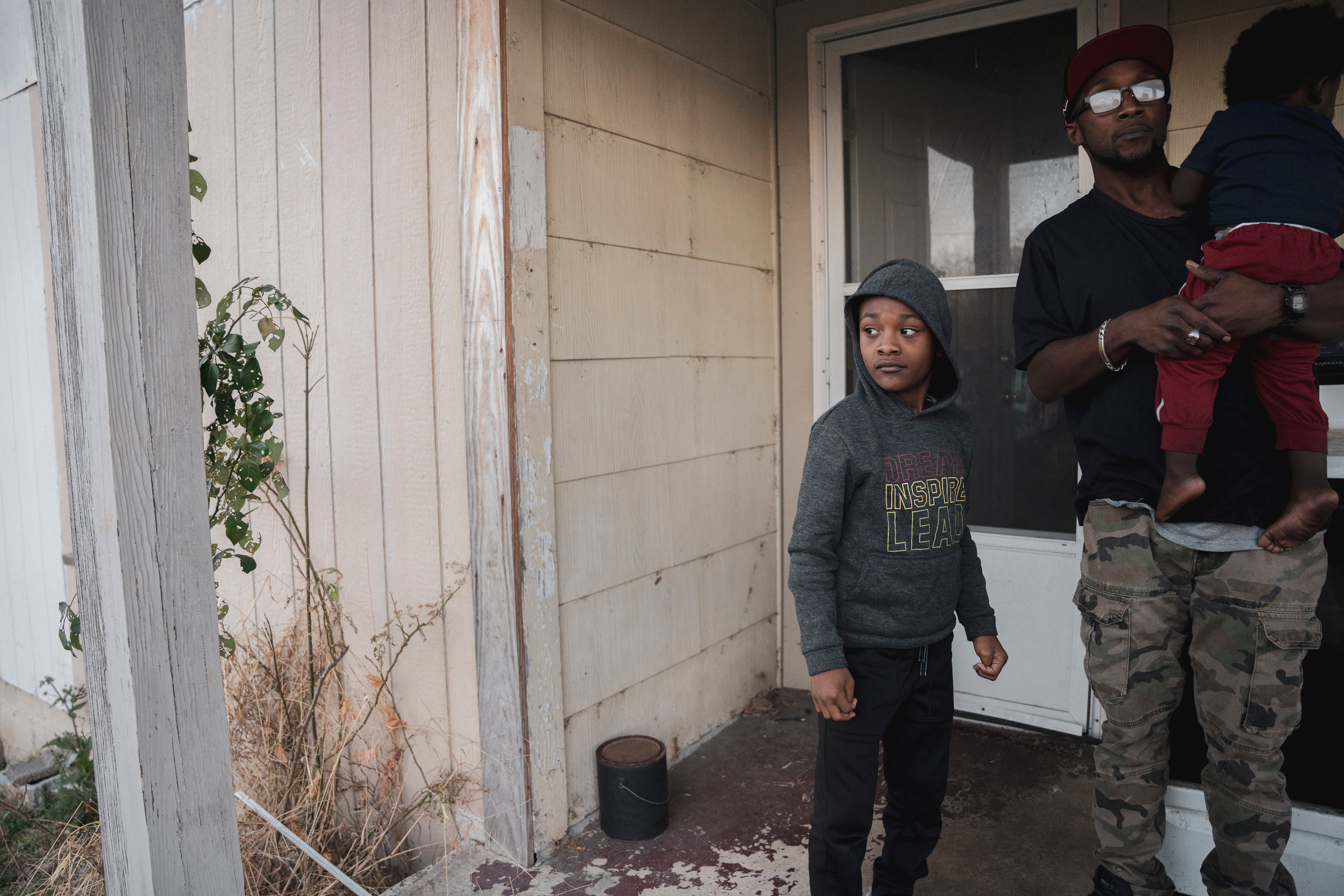 One man and two children stand on a porch near the front entrance to their home.