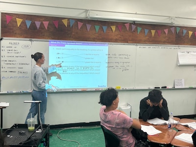 A teacher stands at a lit-up white board in a classroom. She is wearing a light shirt and jeans. Two students sit at their desks, studying. They both have dark hair.