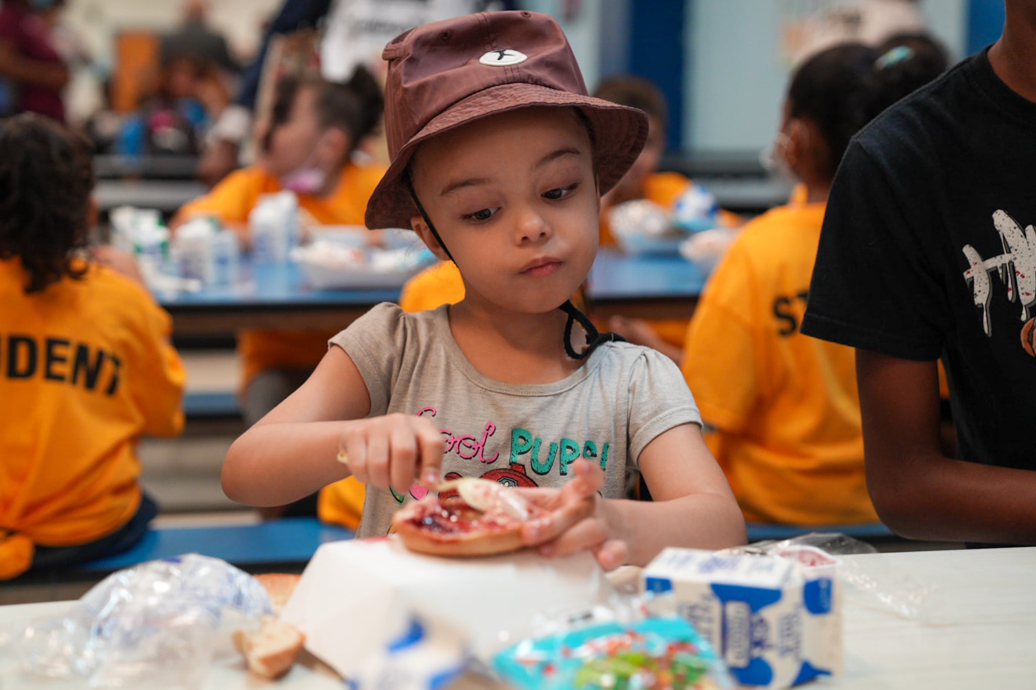 A young girl at a school cafeteria smears cream cheese on a bagel.