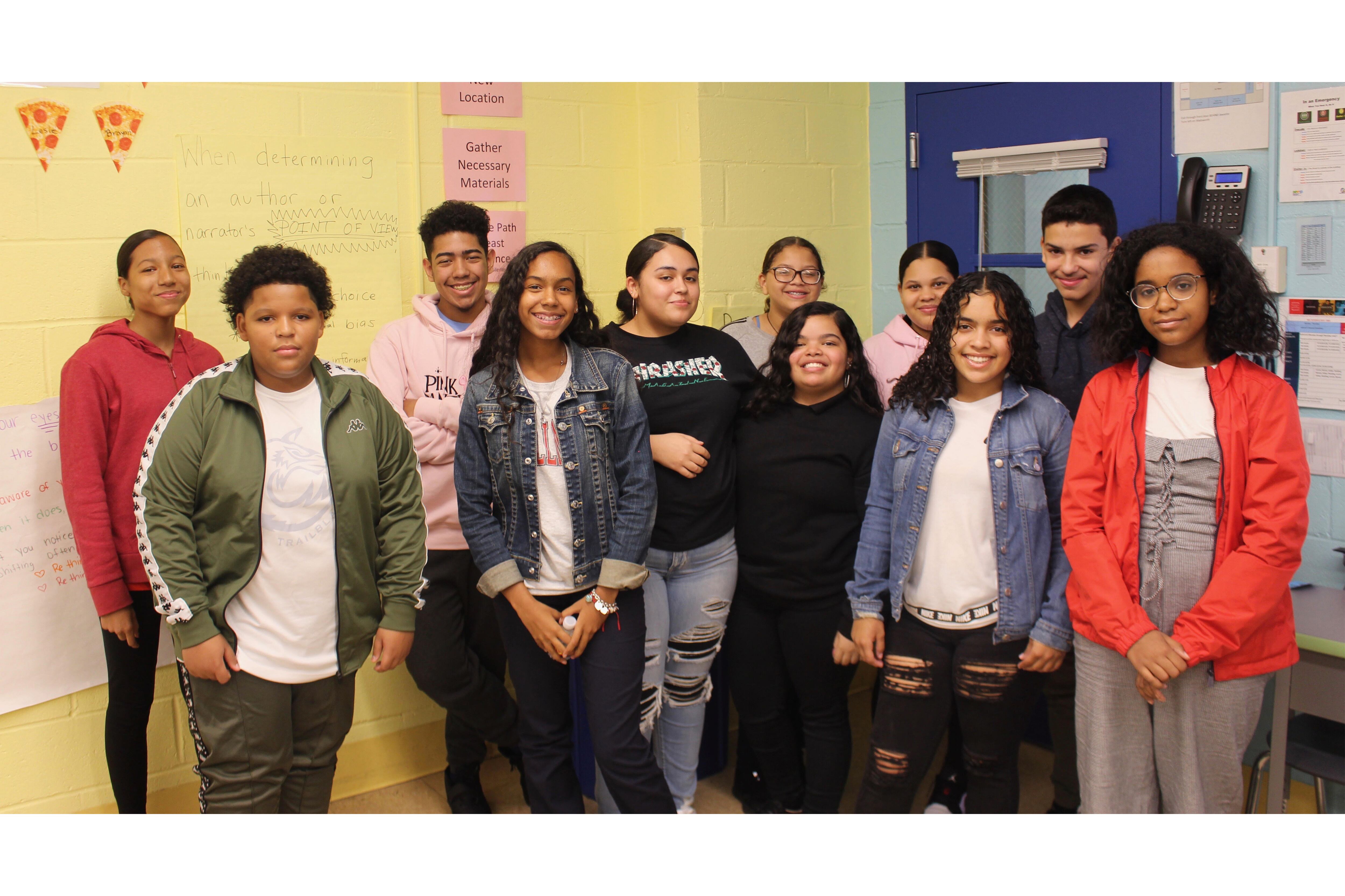 11 students pose for a portrait together in a classroom with yellow walls.